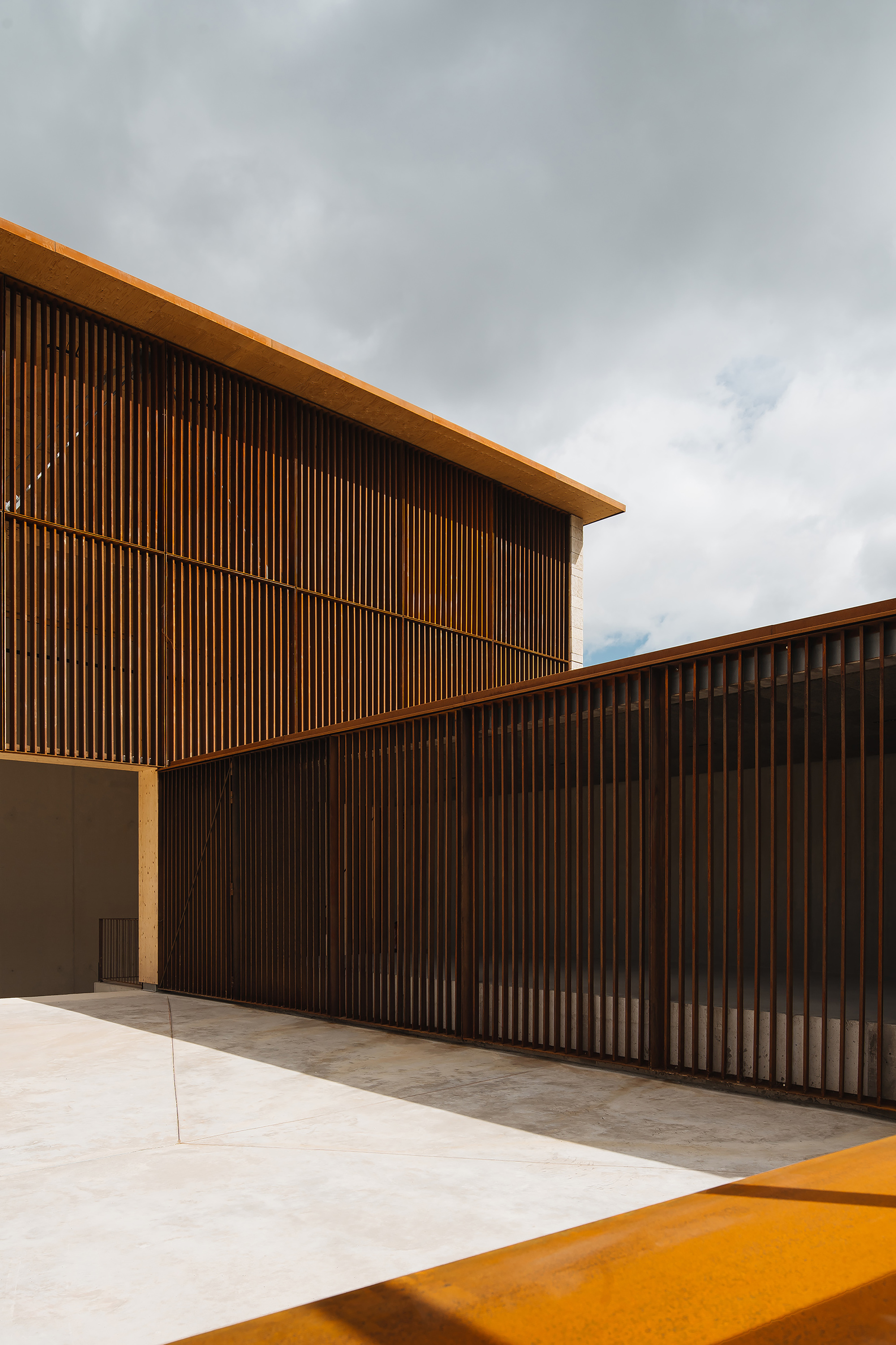 Courtyard with weathered steel slat facades, a pale paved floor and an orange steel edge in the foreground under heavy clouds