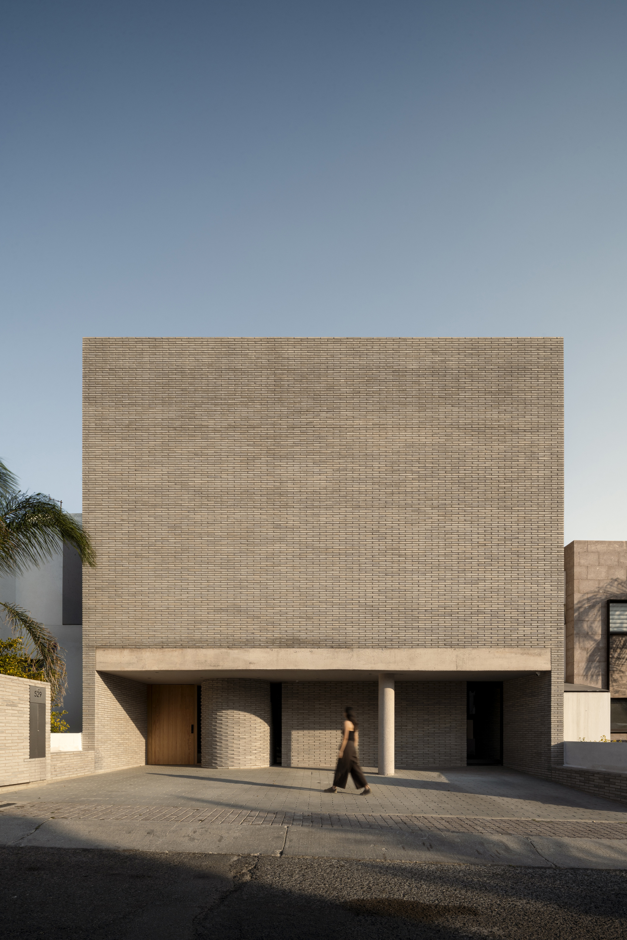 Street facade of a gray brick block raised on a slim concrete column, with a passer-by in late light