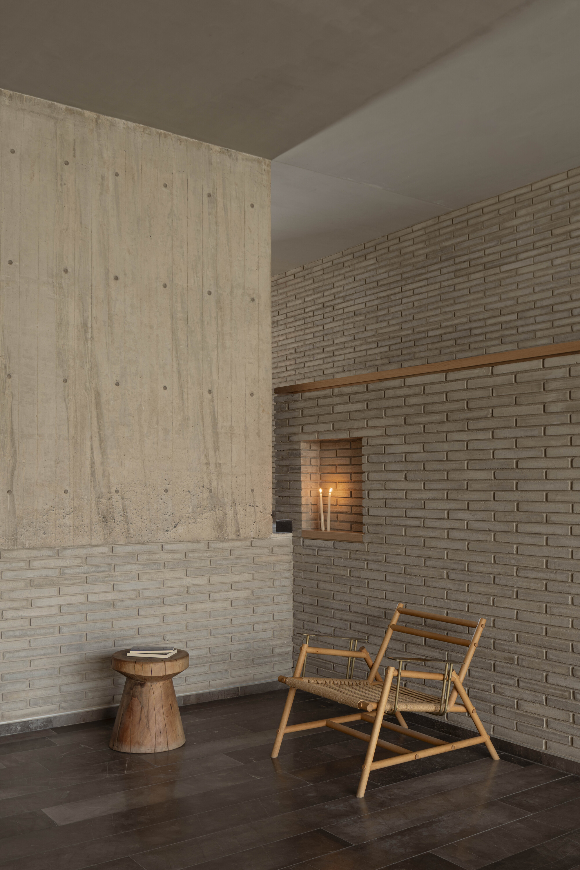 Interior corner of board-marked concrete and slim brick with a lit candle niche, a bamboo chair and a wooden stool