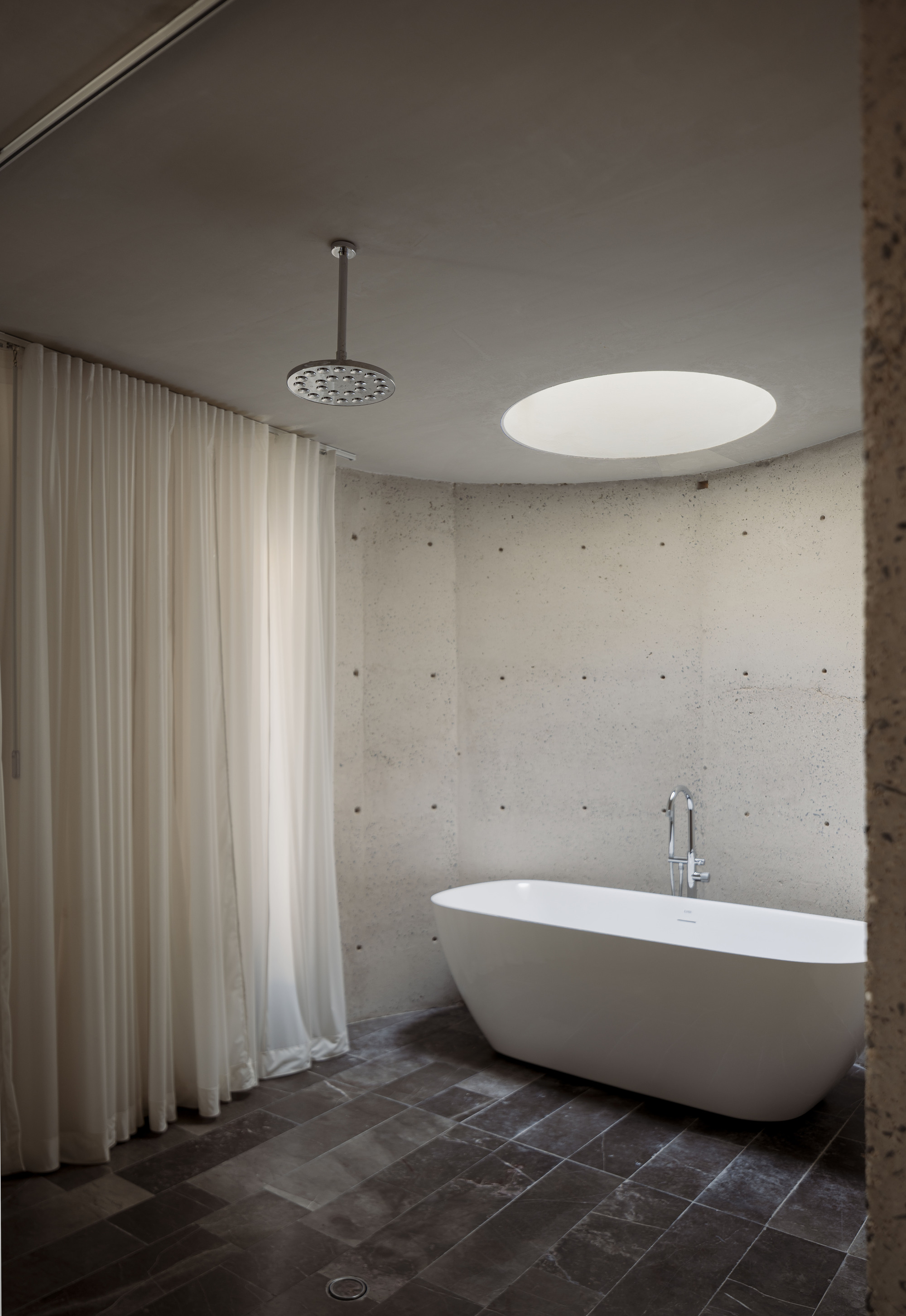 Curved concrete bathroom with a freestanding tub, round skylight, rain showerhead and sheer curtain along dark stone flooring