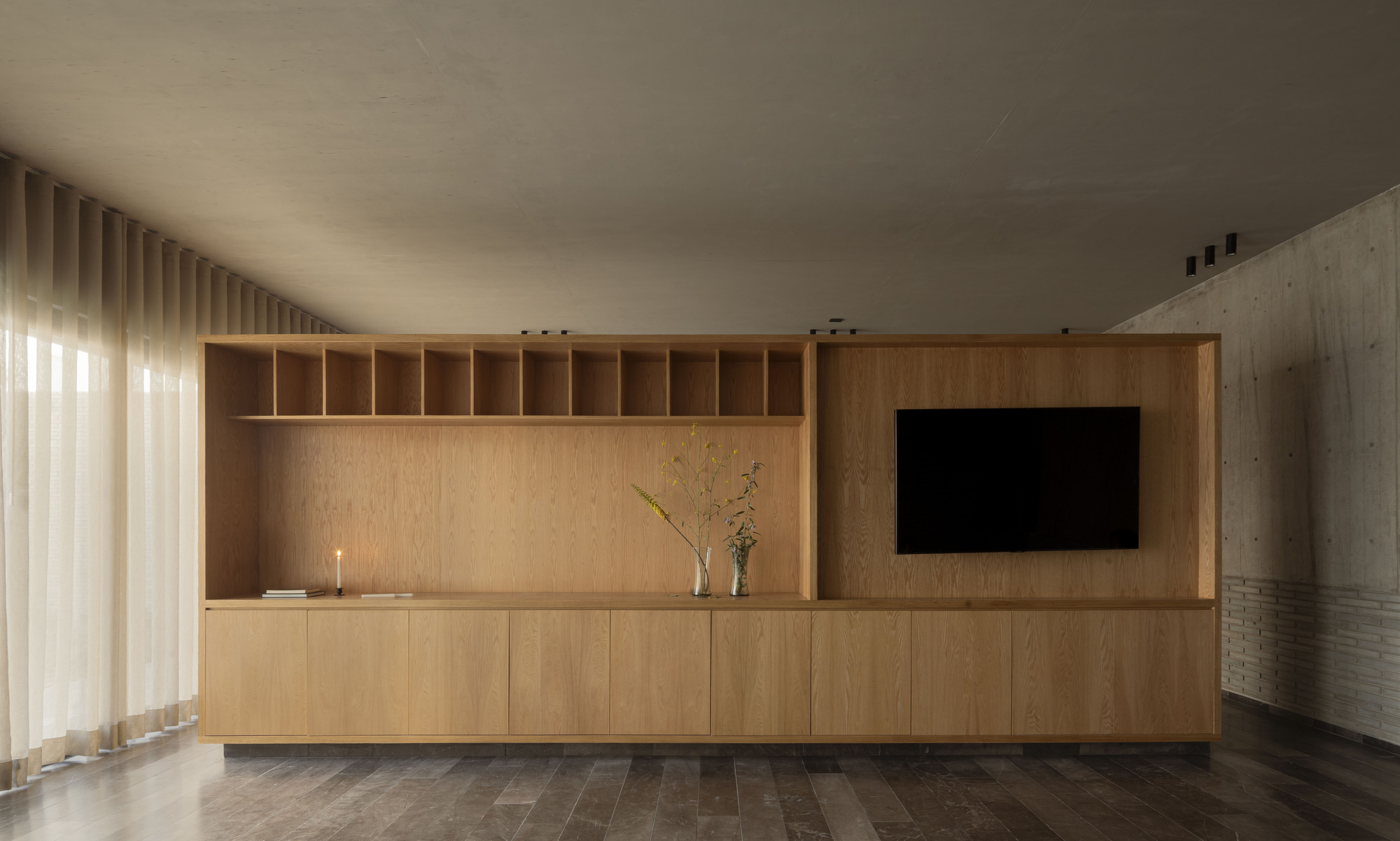 Oak built-in cabinet with open shelves and a recessed television, a vase of dried stems and a candle, beside sheer curtains