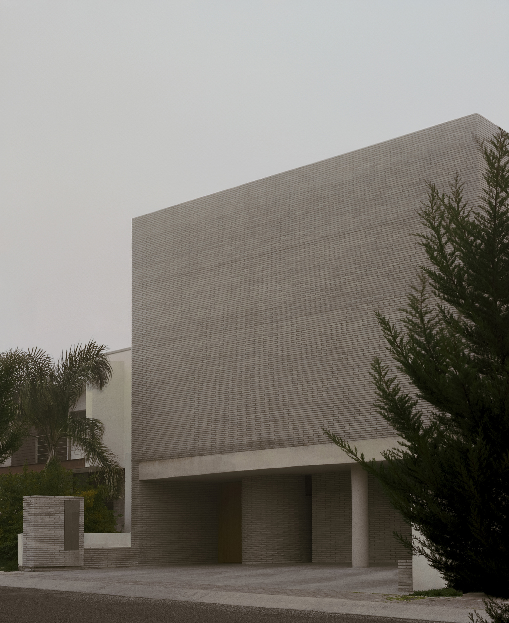 Street view of the long-format brick block cantilevered above a carport, with a palm and a conifer in hazy light