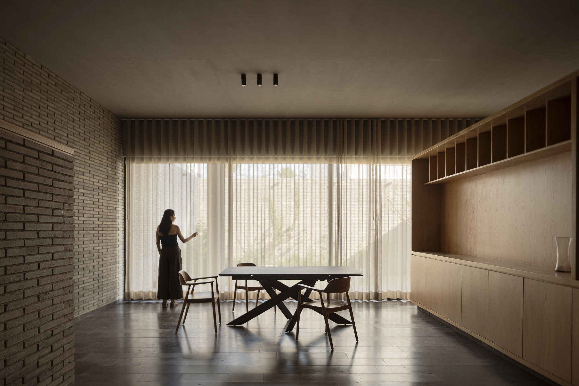 Dining room with a steel-legged table and timber chairs, a woman standing at sheer curtains and oak shelving on the right