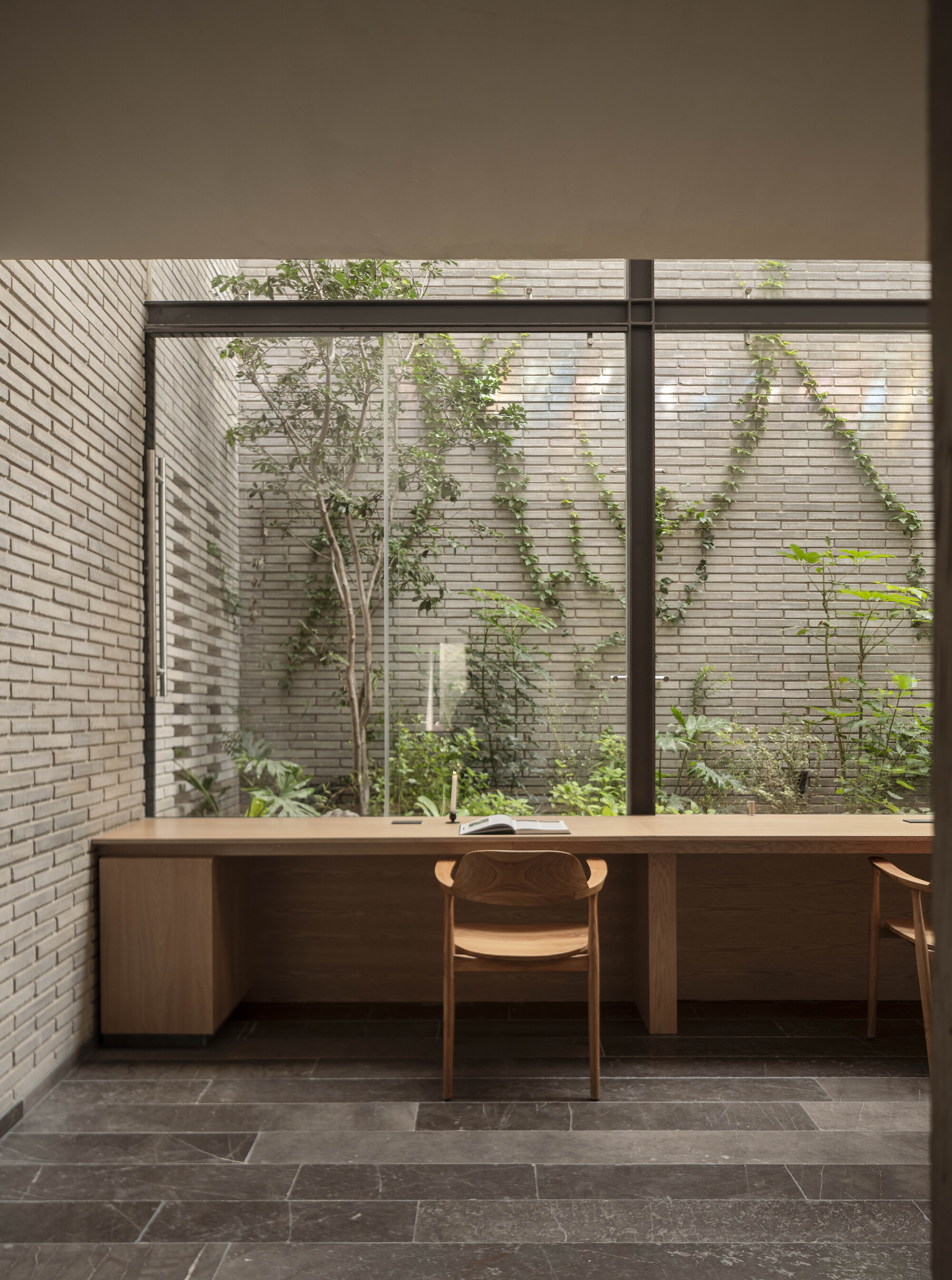 Built-in oak desk and wooden chairs facing a glazed brick courtyard with climbing vines and a slender tree in daylight