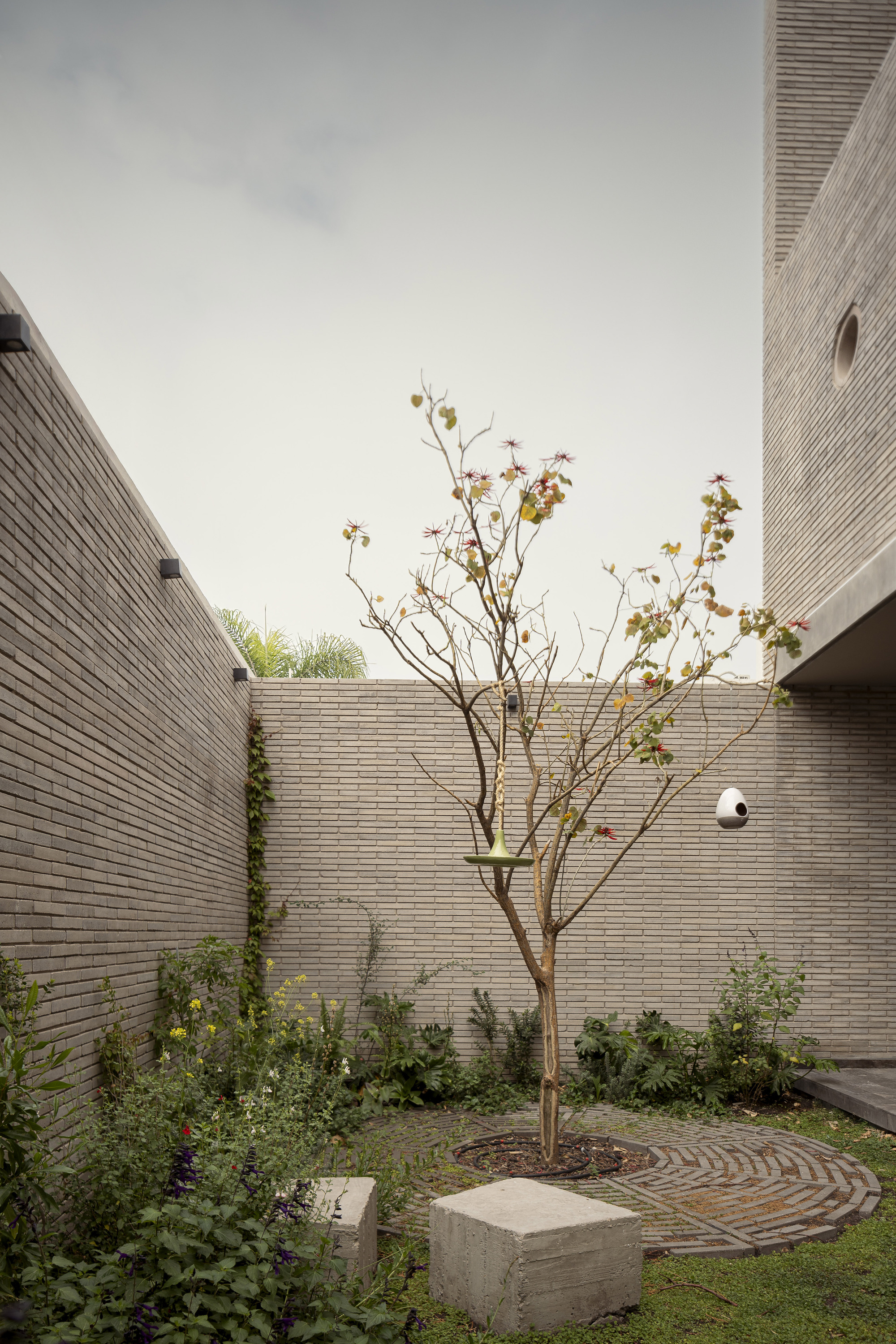 Brick-walled courtyard with a slender flowering tree, circular brick paving and concrete block seats under an overcast sky