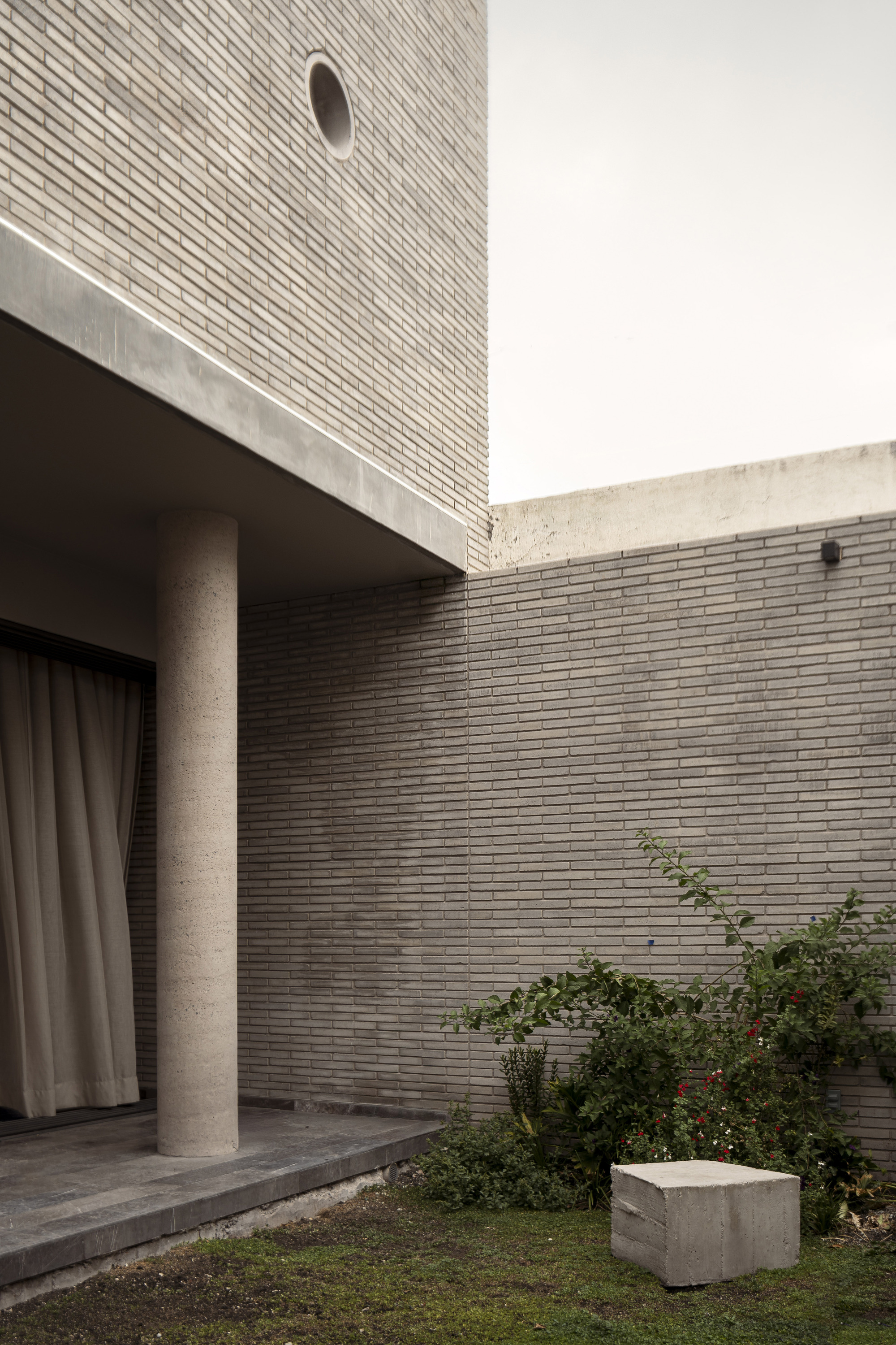 Round concrete column under a cantilevered slab beside a gray brick wall, with a sheer curtain and a flowering shrub