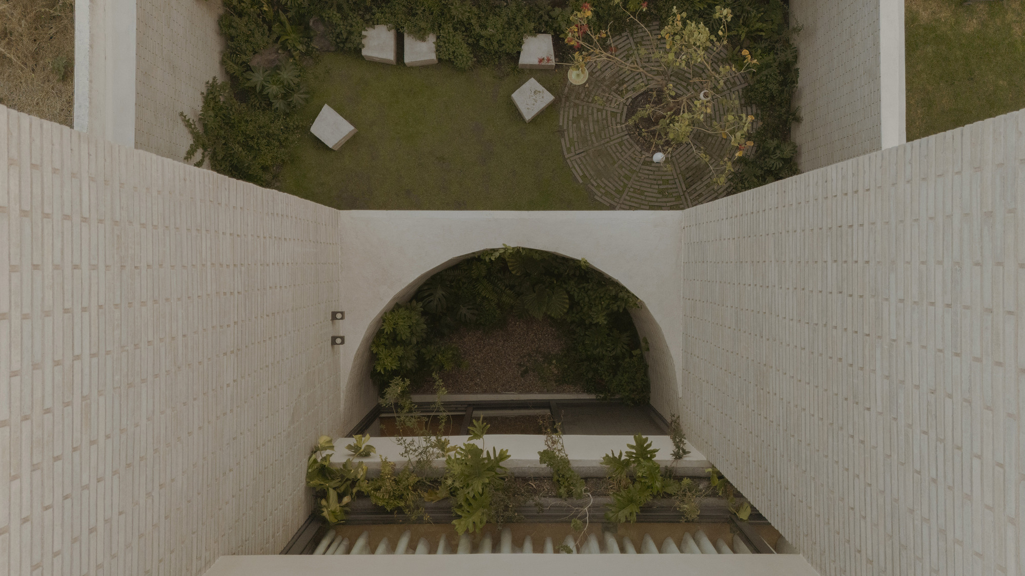 Top-down view of a vaulted brick passage opening onto a lawn with a circular paved tree bed and concrete blocks