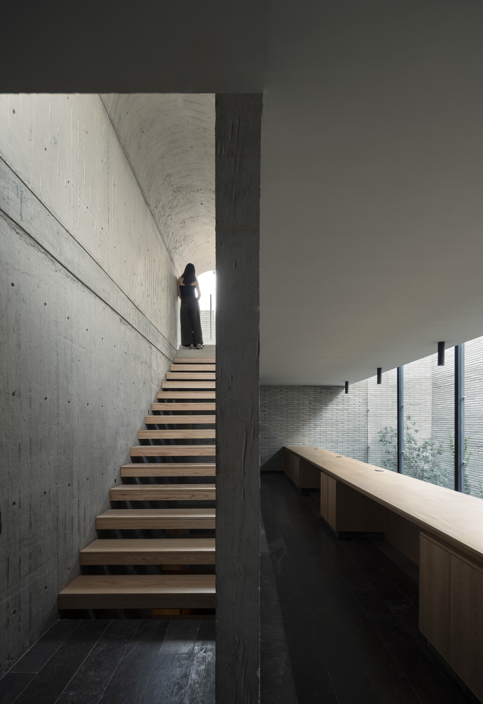 Figure standing at the top of a timber stair under a concrete barrel vault, beside a long built-in wooden desk