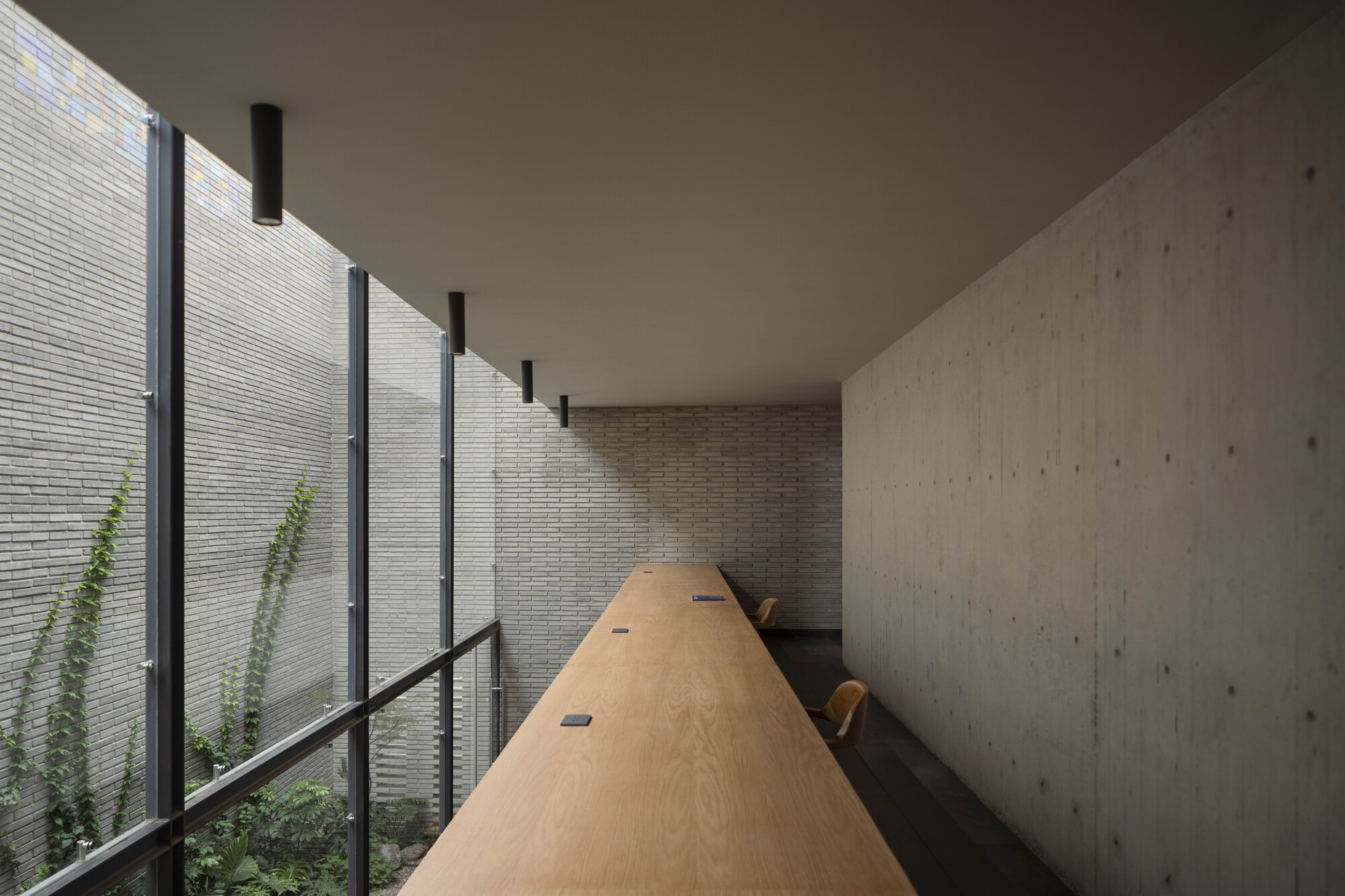 Long timber table running beside a glazed wall facing a brick light well with climbing vines, concrete wall at right
