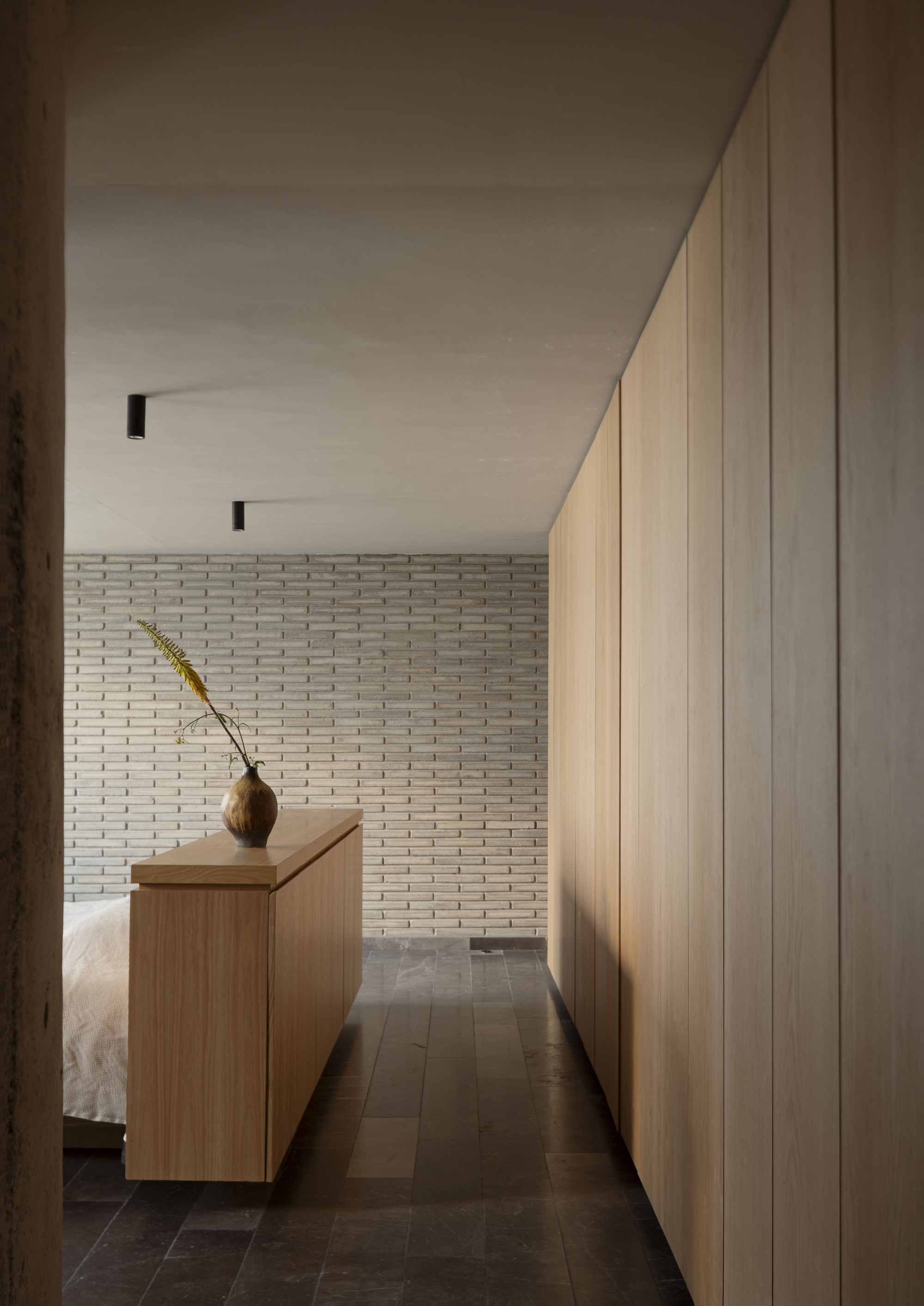 Oak storage wall and low timber cabinet with a vase of dried grass, facing a pale brick wall in soft light