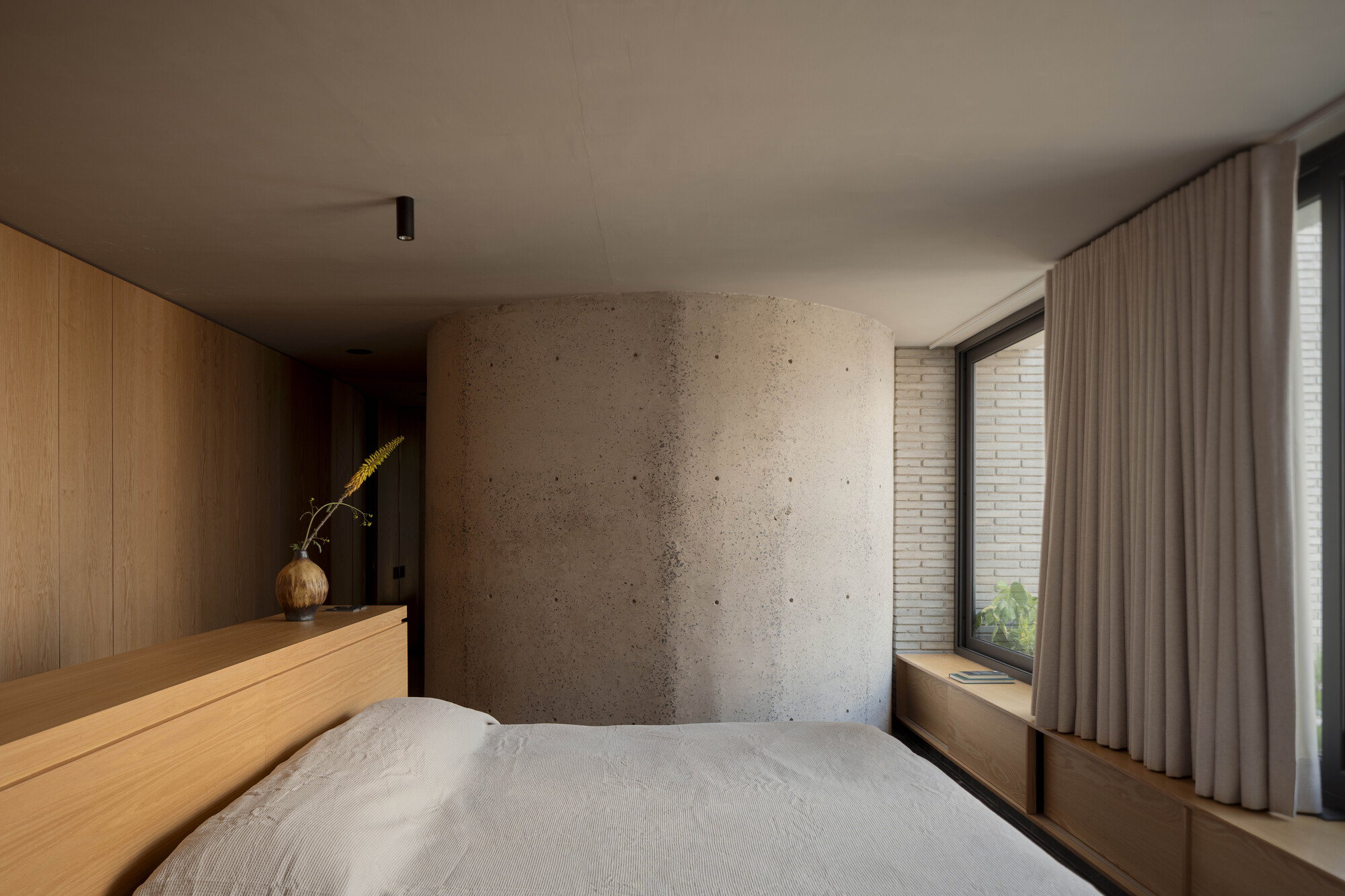 Bedroom with a rough curved concrete column, oak headboard unit and linen curtain beside a window seat in dim daylight
