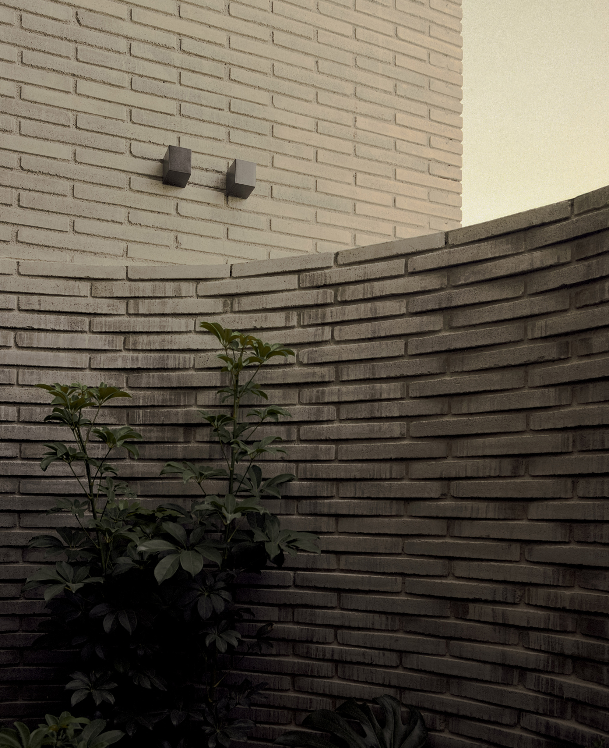 Curved brick garden wall with a leafy shrub in the foreground and two small wall lights on the brick above at dusk