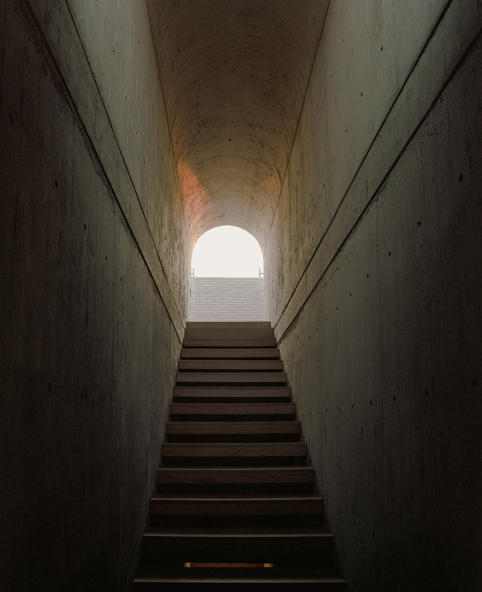 Narrow concrete stairwell with timber treads rising to a barrel-vaulted opening that glows with bright daylight and a brick wall beyond