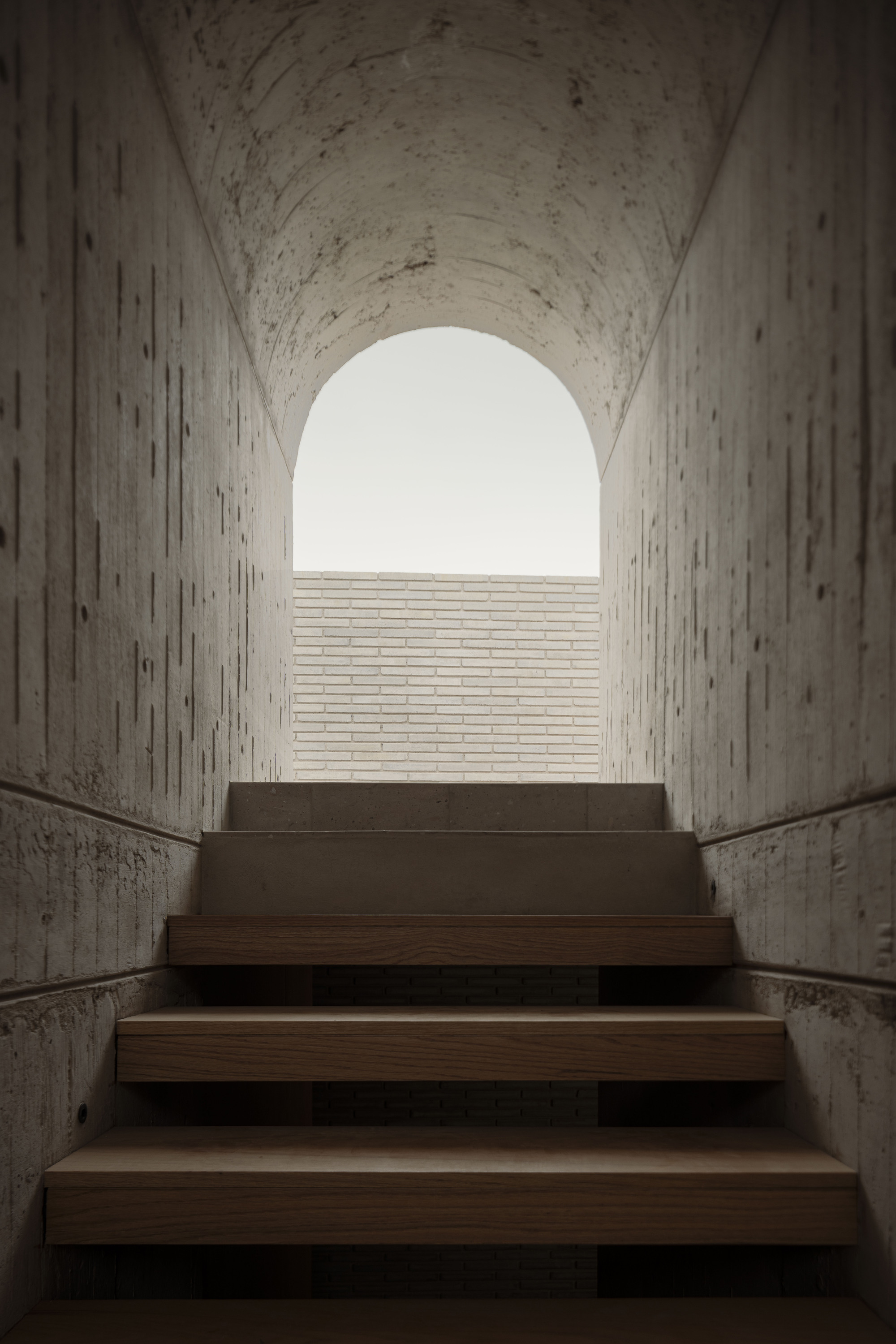 Board-formed concrete stairwell with oak treads rising toward an arched opening and pale brick wall under overcast sky
