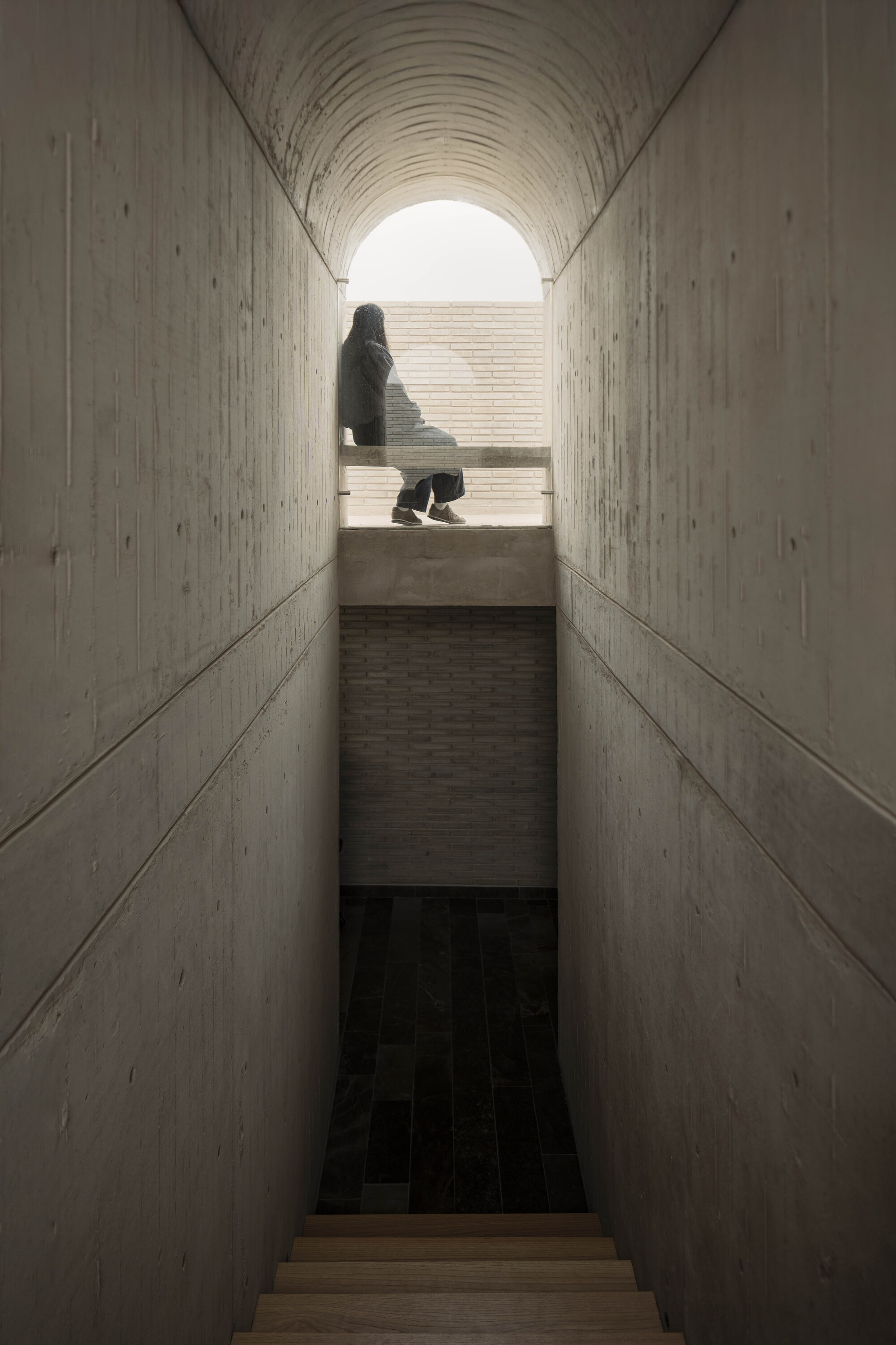 Narrow concrete stair passage with a seated figure silhouetted in the arched opening above, dark stone floor below