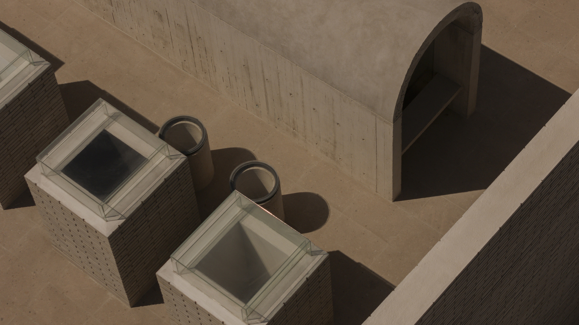 Rooftop view of glazed skylights on brick plinths beside a concrete barrel vault, casting long shadows in warm light
