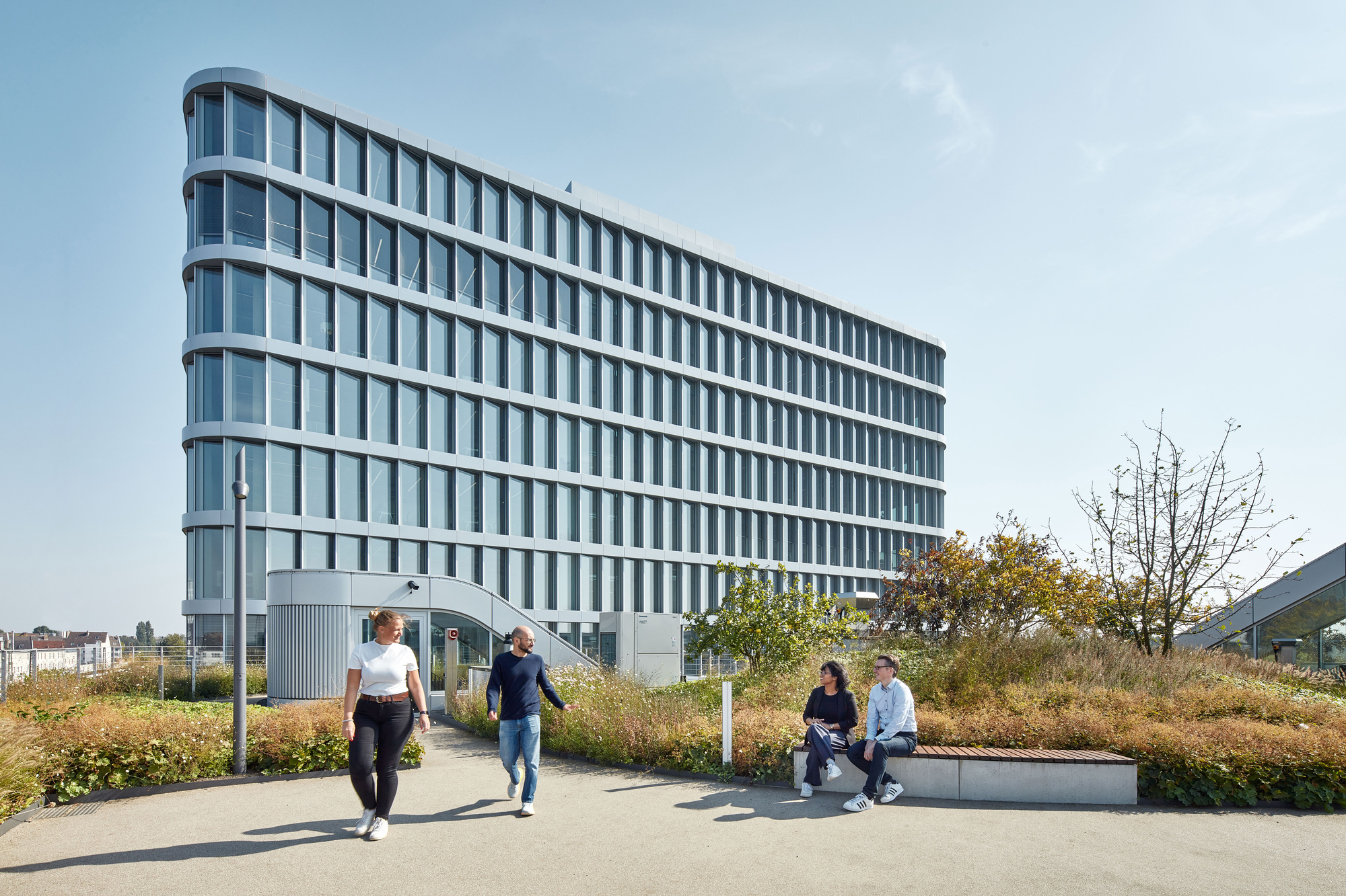 Tall curved office tower with horizontal bands of glazing, seen from a roof garden where people walk and sit on a bench