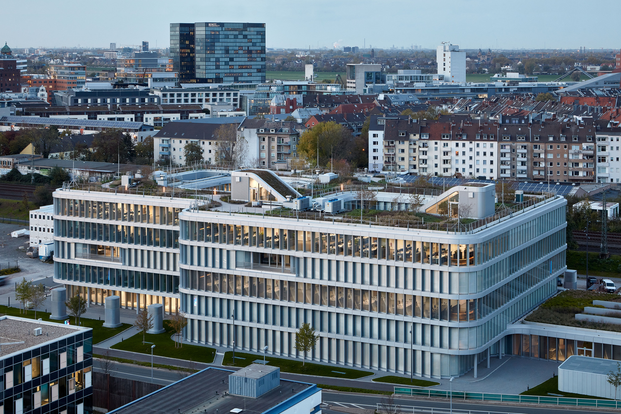 Elevated view of the curved office facade with fine vertical fins and a planted roof terrace at dusk