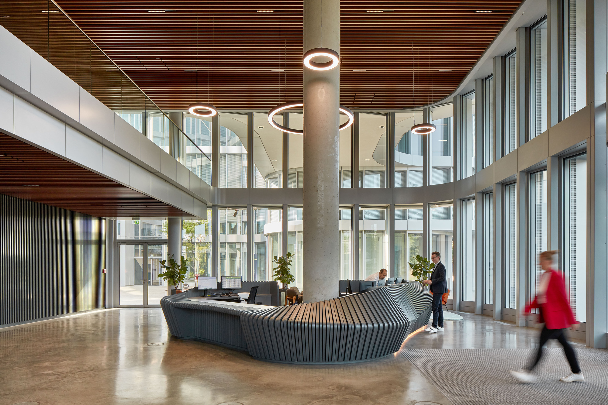 Lobby with a sculpted fluted reception desk around a concrete column and a timber slat ceiling, a person in red walking past