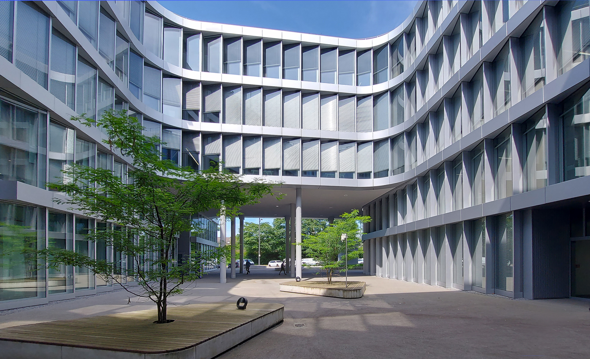 Courtyard between curved glass facades, with wooden planter benches and young trees beneath a covered passage in bright sunlight