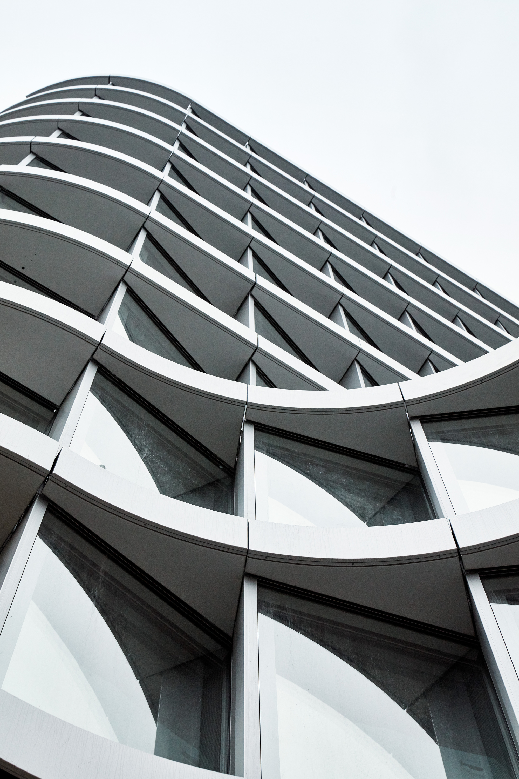 Upward view of the undulating facade with curved white fins and angled glass panes against an overcast sky