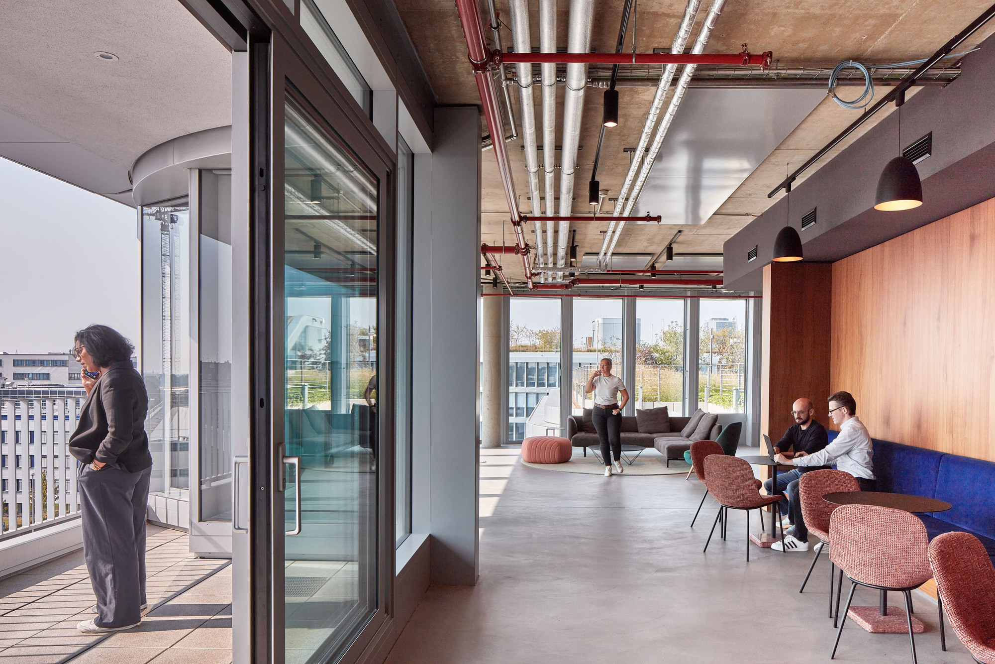 Open office lounge with sliding glass doors, exposed red pipes and a timber-clad wall, a woman standing on the balcony in daylight