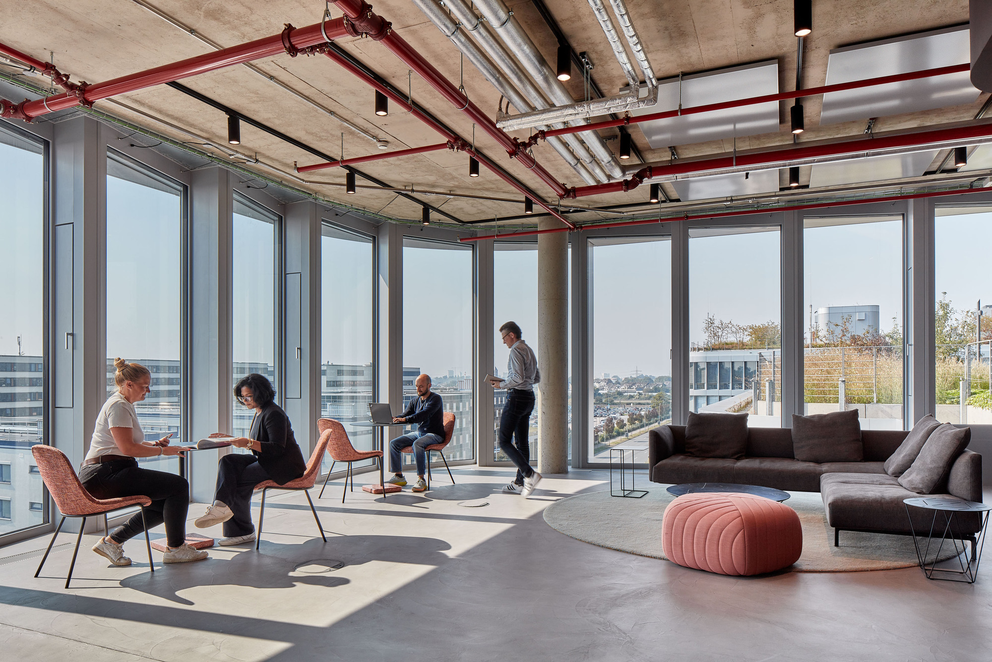 Corner workspace with floor-to-ceiling glazing, exposed red services overhead and a pink pouf beside a grey sofa in strong sunlight