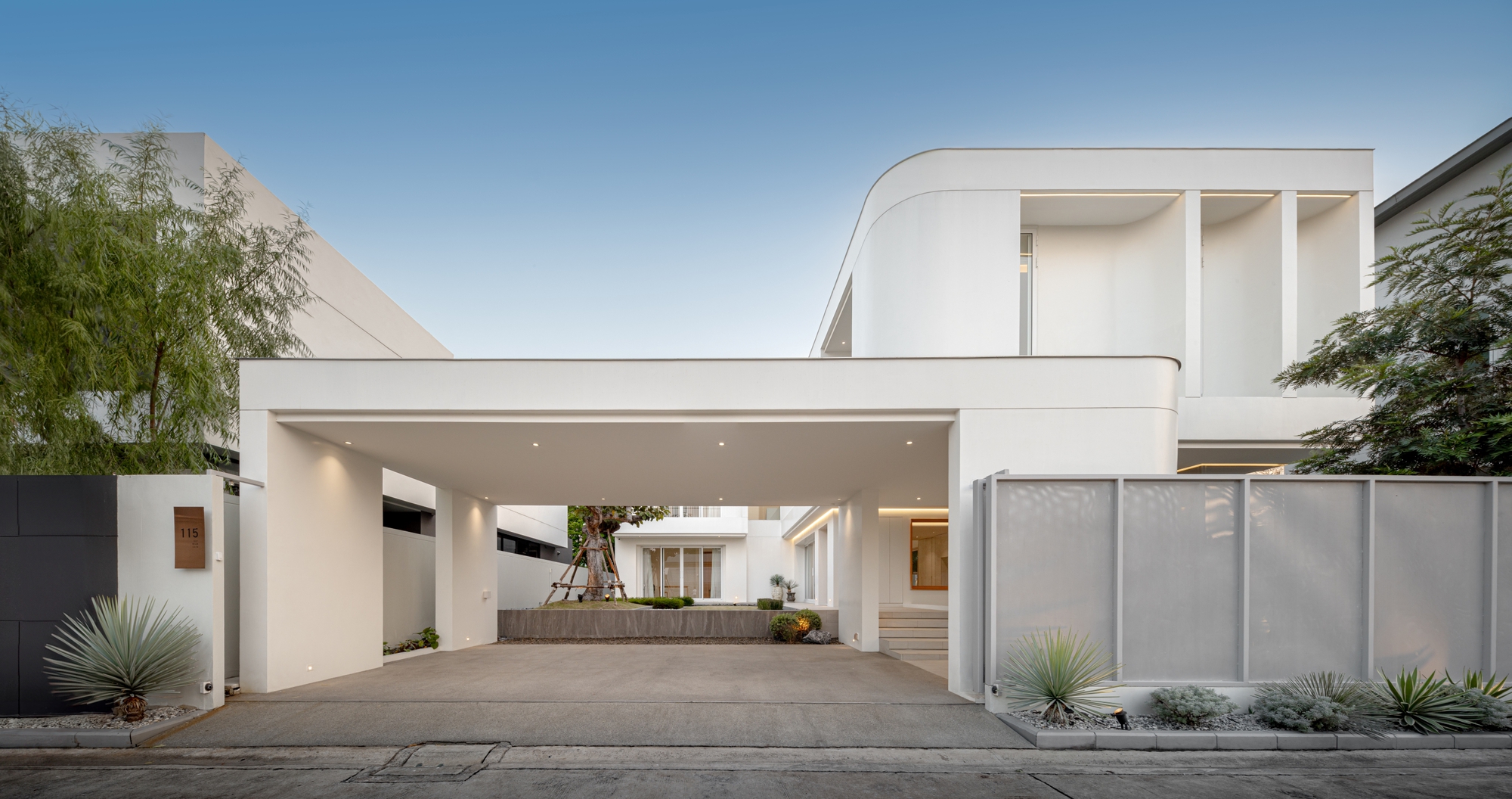 White street facade with a deep carport canopy, curved upper volume and perforated metal screen fence under a clear dusk sky