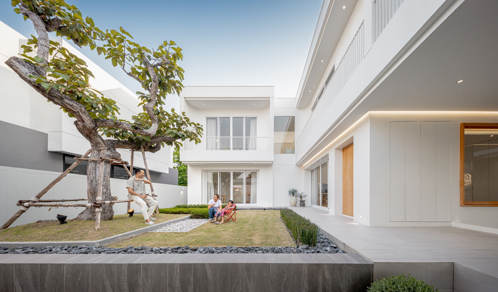Family sitting on the courtyard lawn beside a propped, staked tree, with pebble beds and a white two-storey wing behind