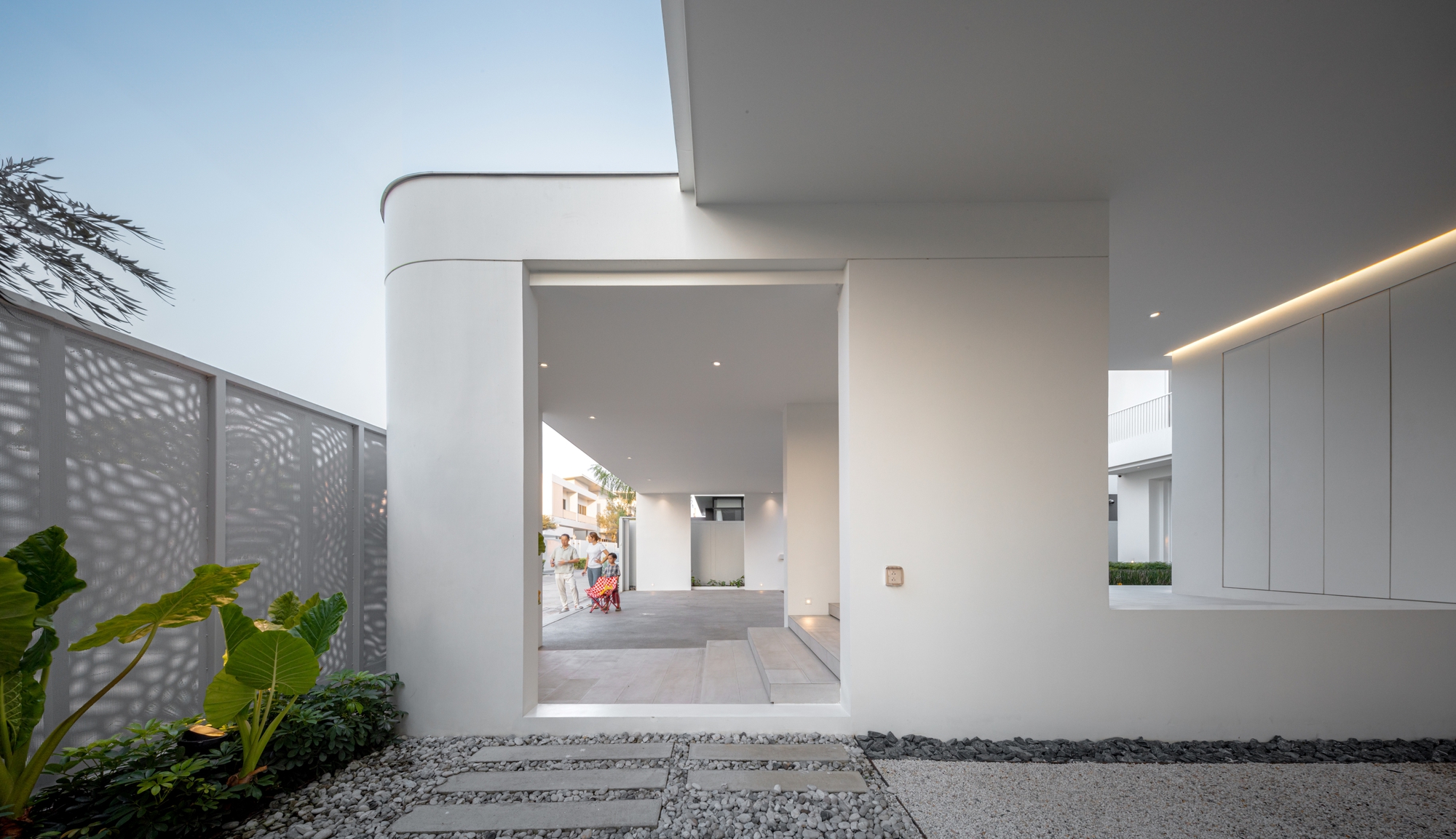 Framed opening through a white curved wall toward the carport, with stepping stones, gravel and broad-leaf plants beside a perforated screen