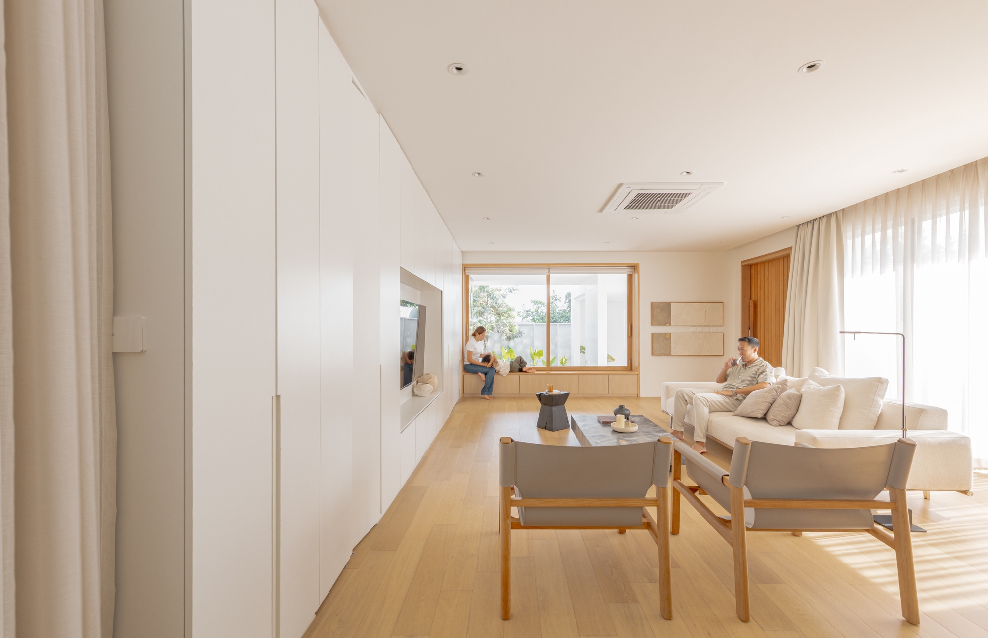 Living room with a long white cabinet wall, oak floor and leather-slung chairs, sunlight falling through sheer curtains as two people relax
