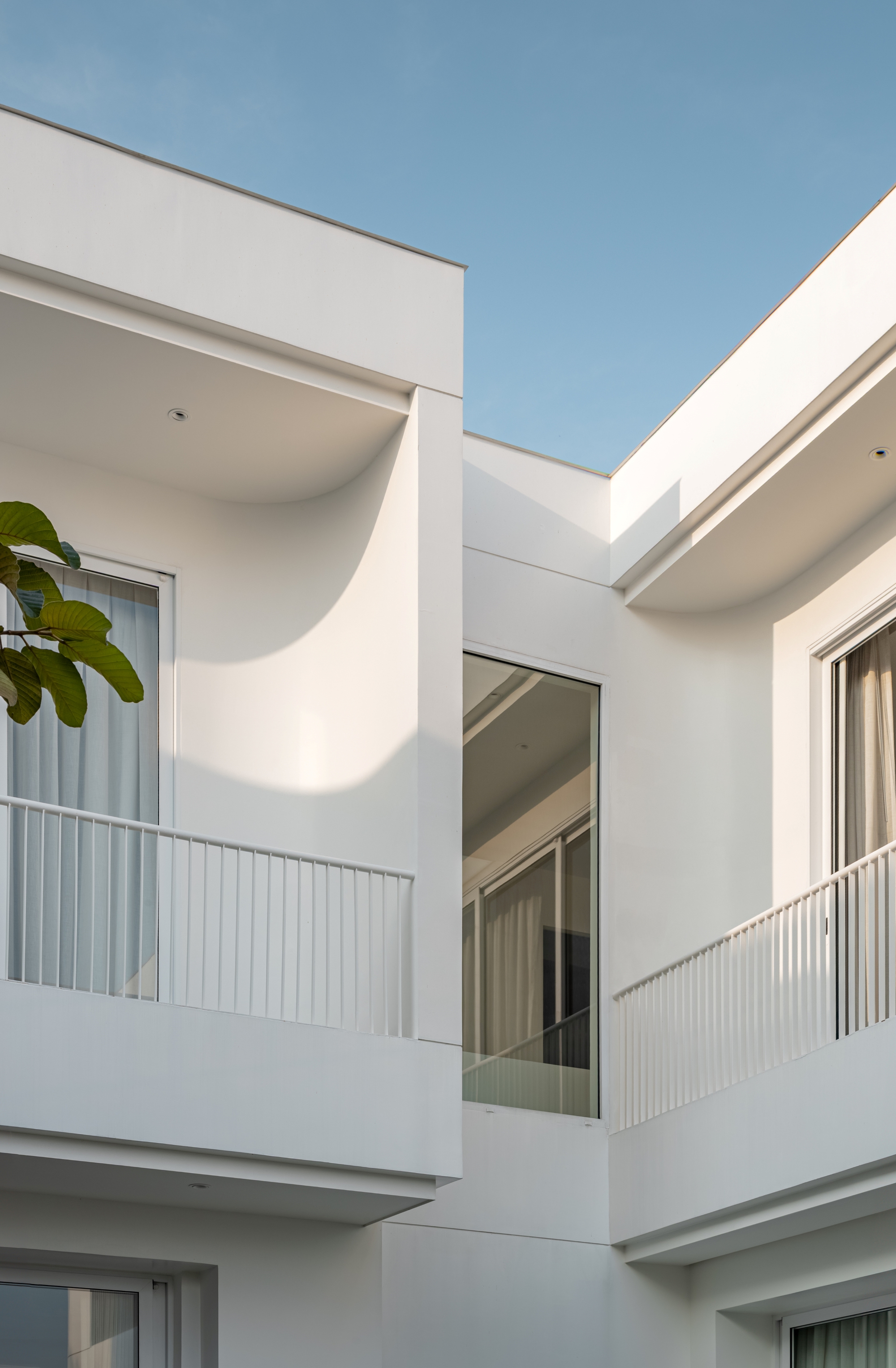 Upper-floor balconies with white balustrades and a curved soffit, a tall glass panel between them and a leafy branch against blue sky