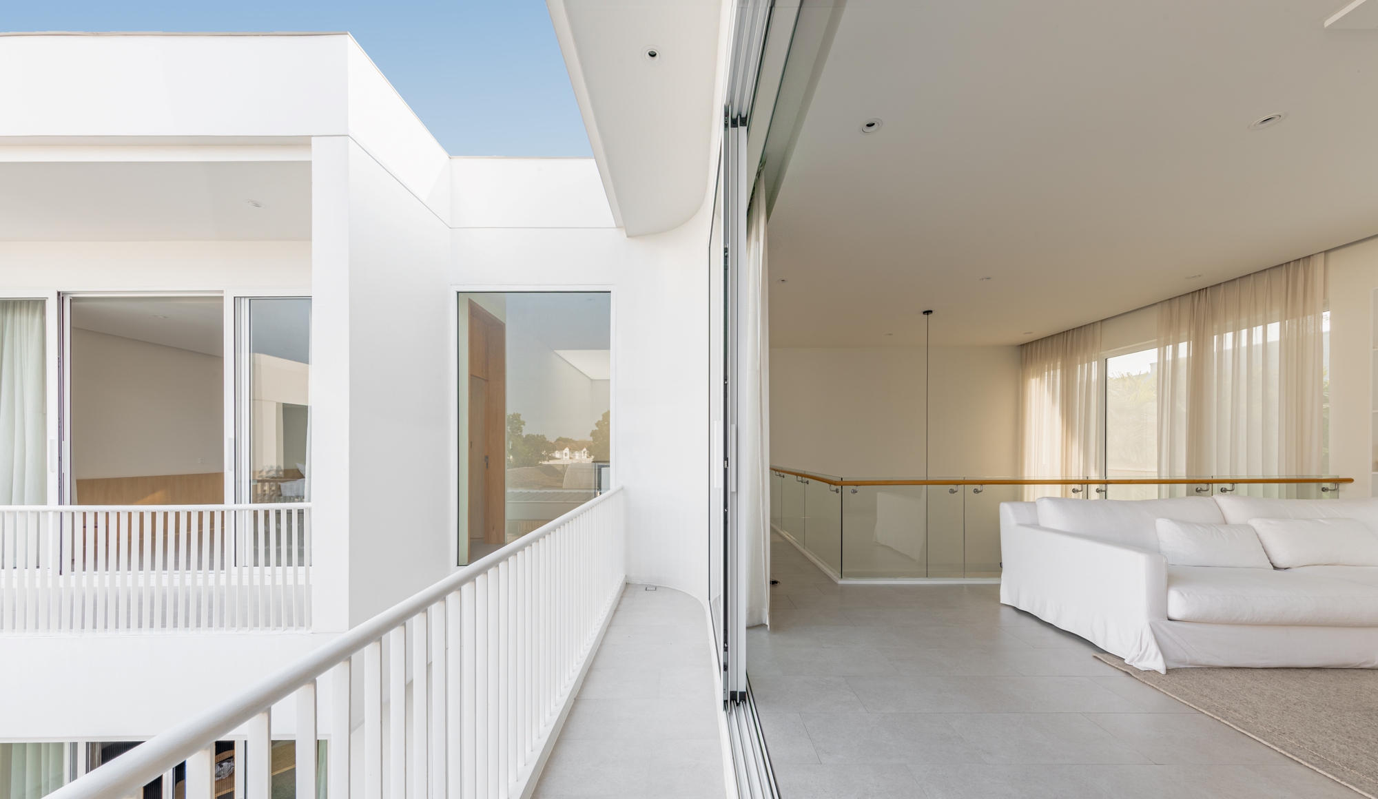 Upper-level balcony with white railing beside a sliding glass wall, opening to a white sofa and glass balustrade in warm afternoon light