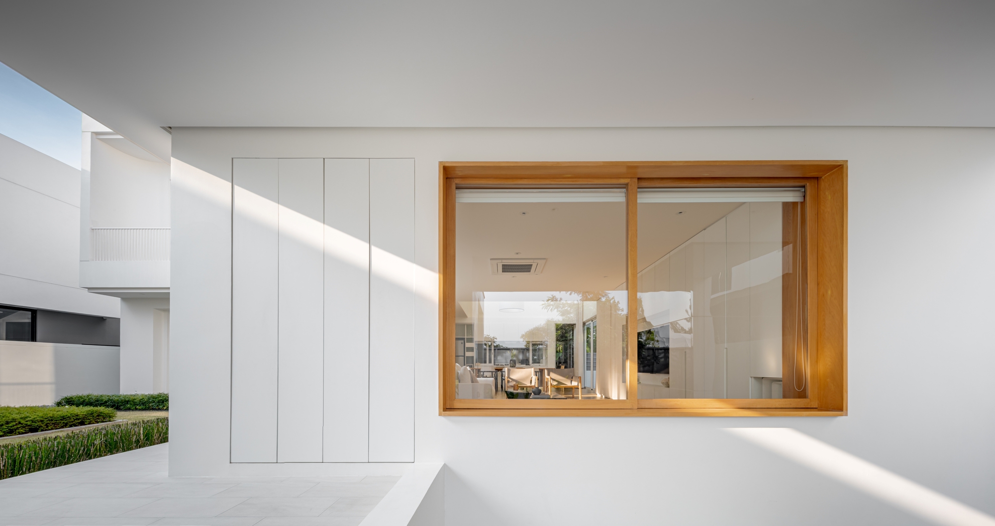 White exterior wall with a timber-framed sliding window and tall flush panels, crossed by diagonal sunlight and a clipped hedge