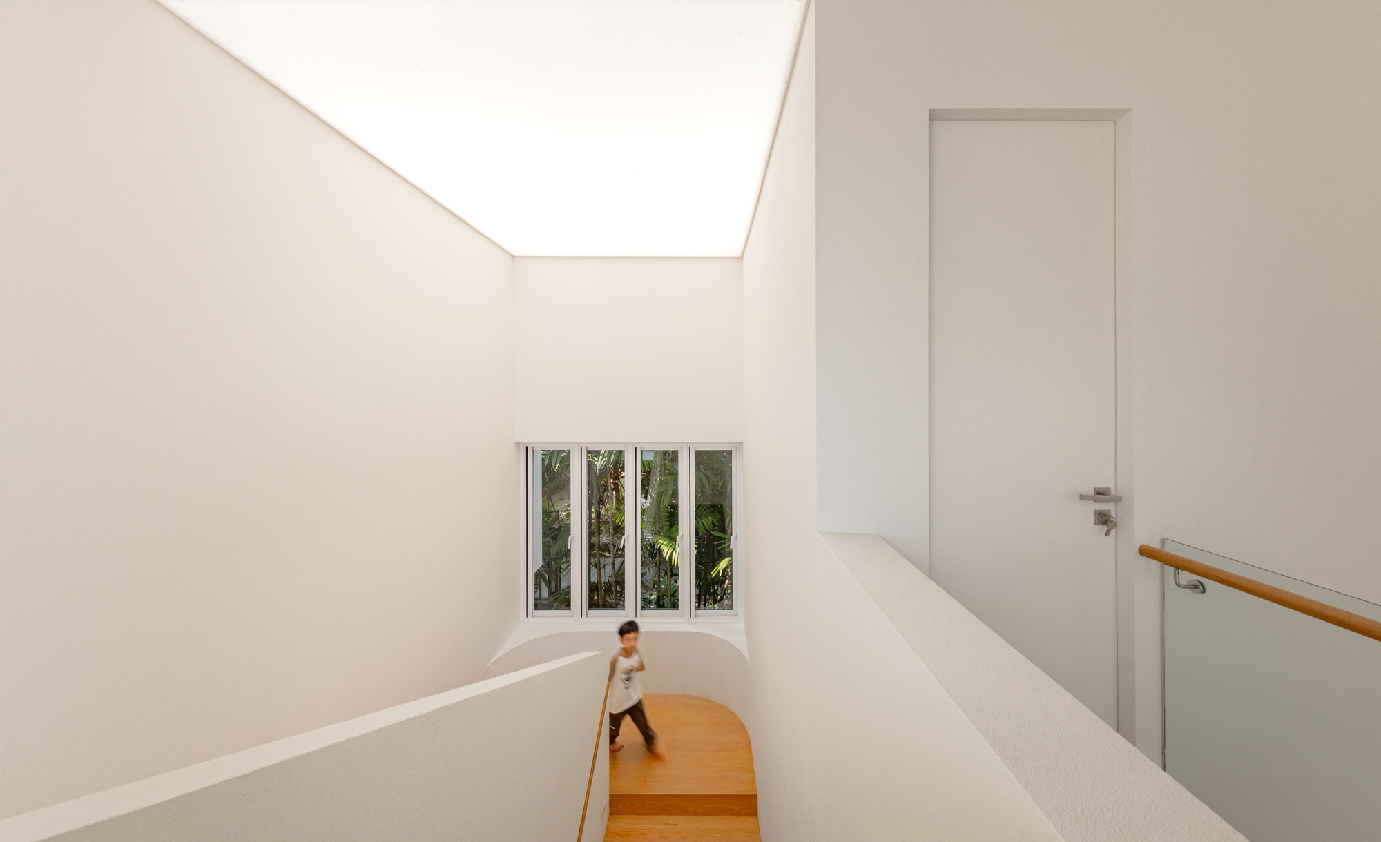 Stairwell with a glowing ceiling panel and a curved landing, a child running down past a window onto palm foliage