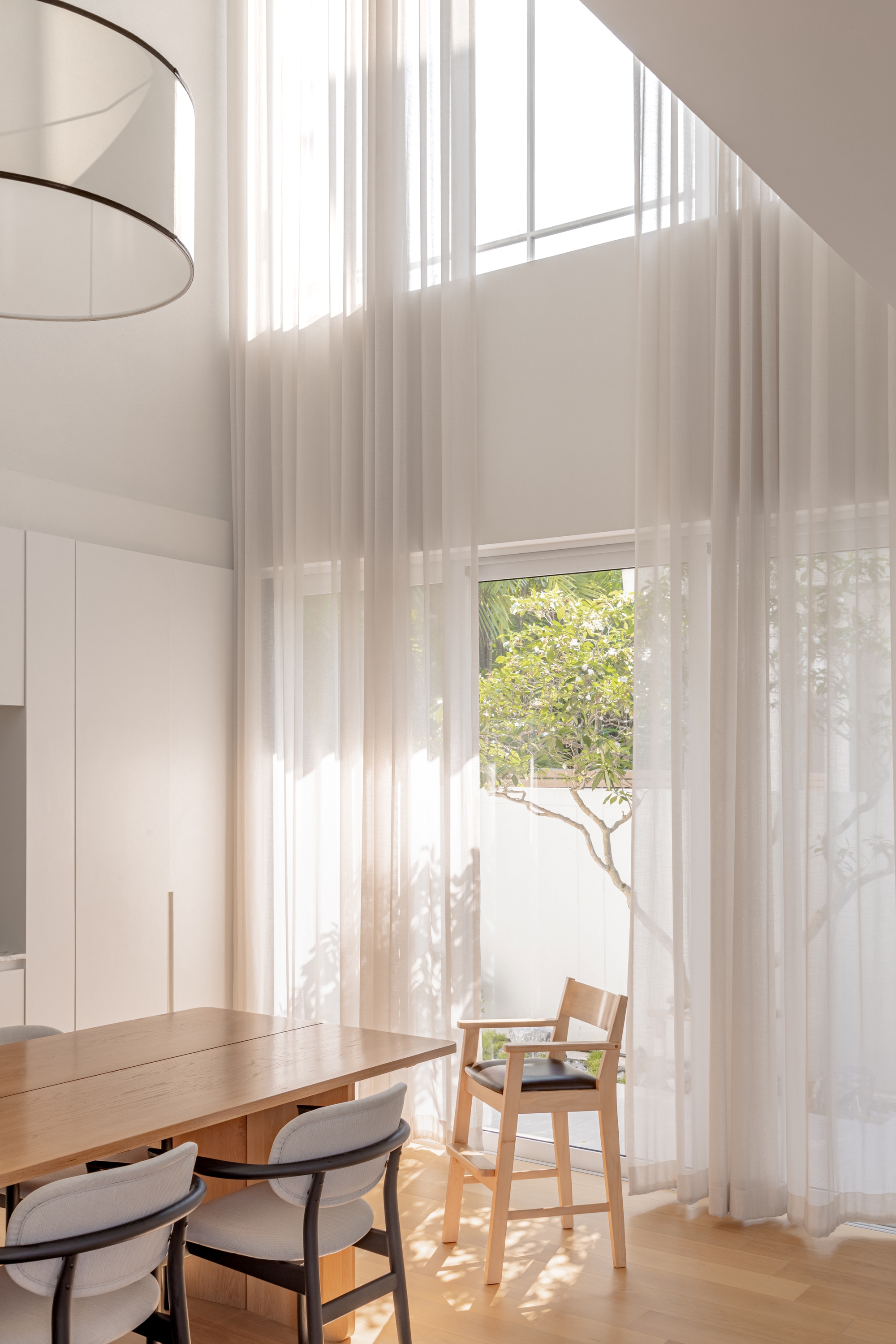 Floor-to-ceiling sheer curtains beside a timber dining table and chairs, with sunlight and a tree outside the tall window