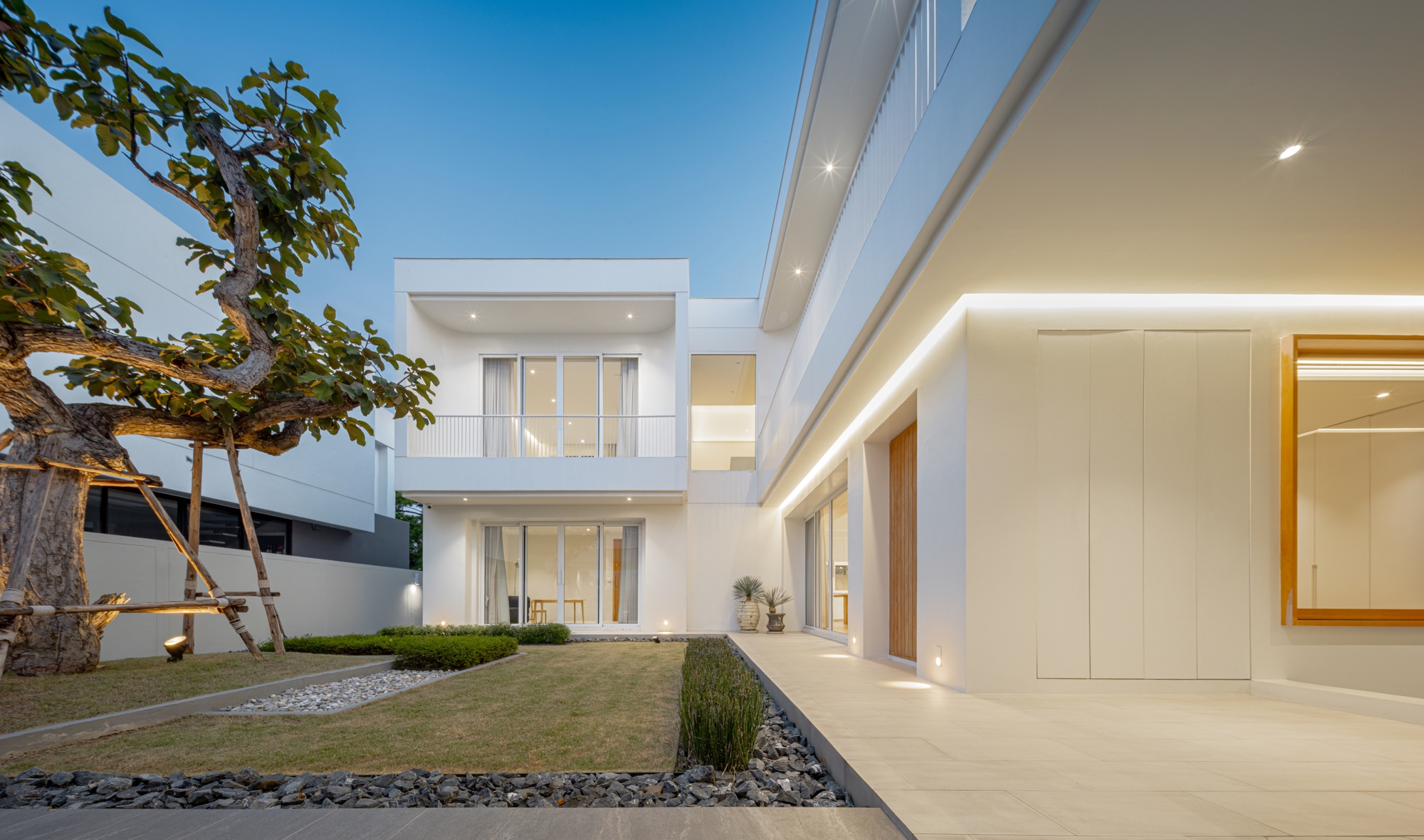 White two-storey house at dusk with a gnarled propped tree, gravel-edged lawn and a cove-lit canopy over the entry