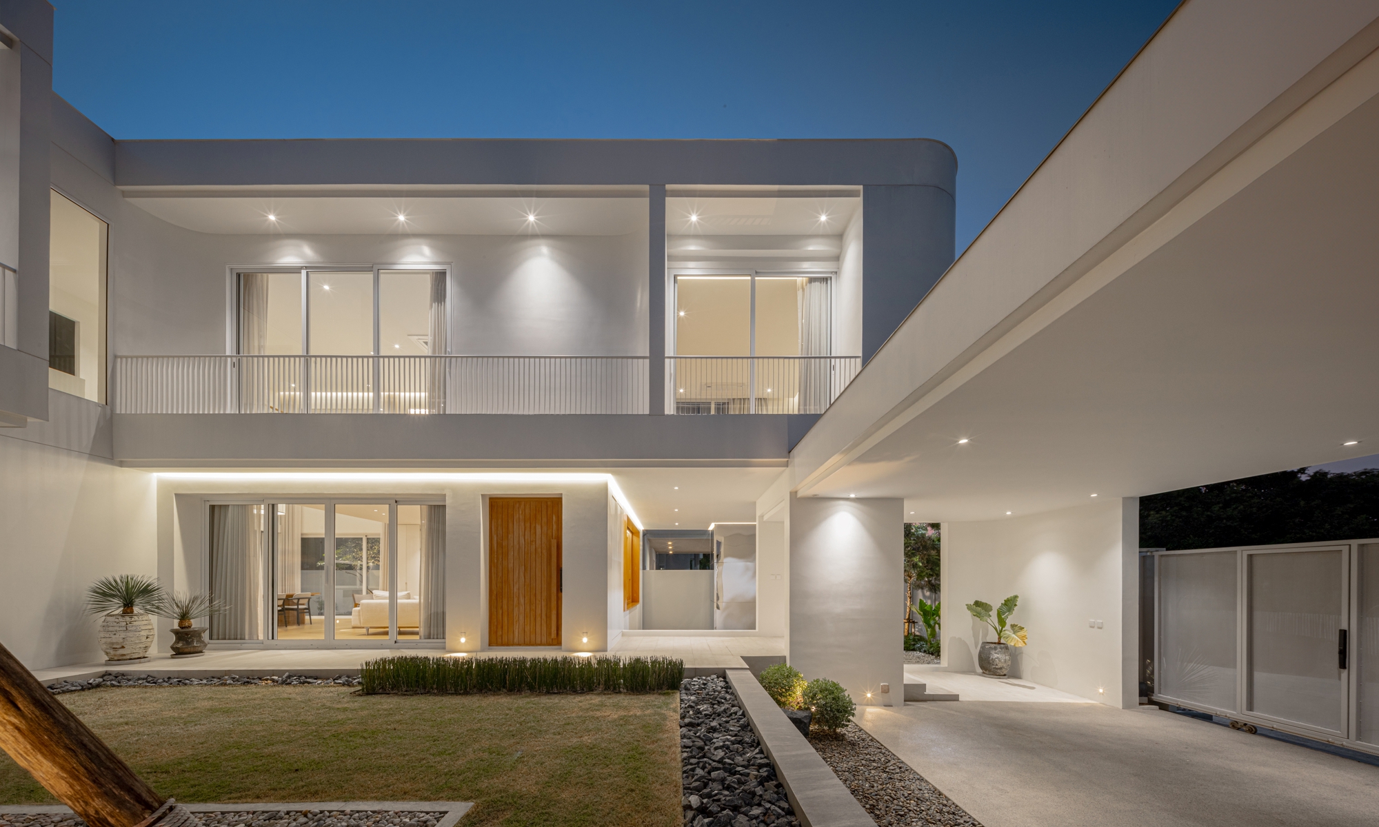 Courtyard elevation at dusk showing a timber front door, upper balcony with slim railings and a carport canopy beside the lawn