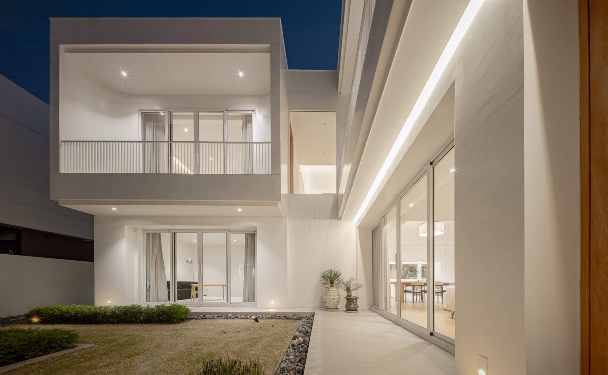 Side terrace at night with potted plants, glass sliding doors and a continuous light strip along the white wall