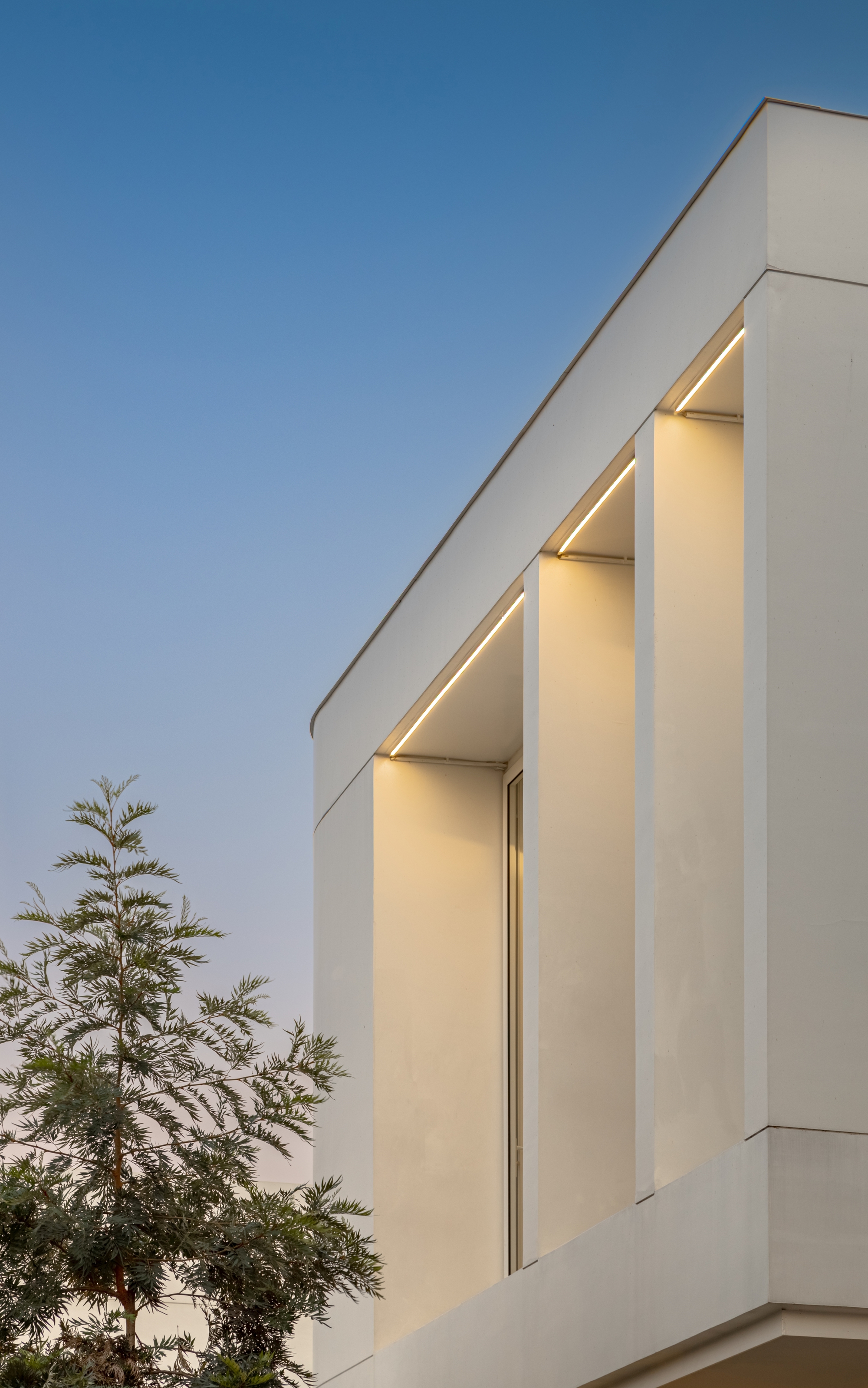 Corner of the upper volume with recessed vertical fins lit by linear lights against a fading blue sky and a feathery tree