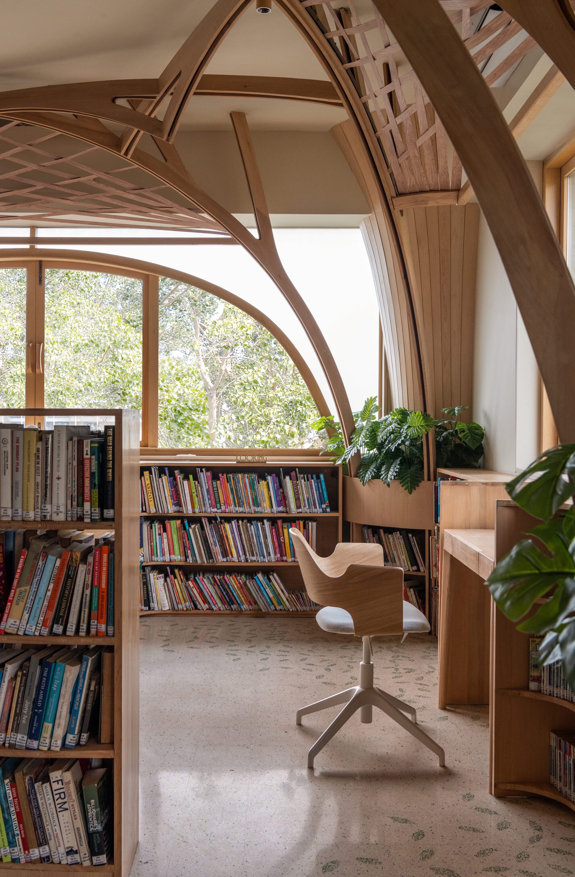 Swivel chair beside a bookshelf, facing an arched window with tree canopy beyond and a lattice timber ceiling above
