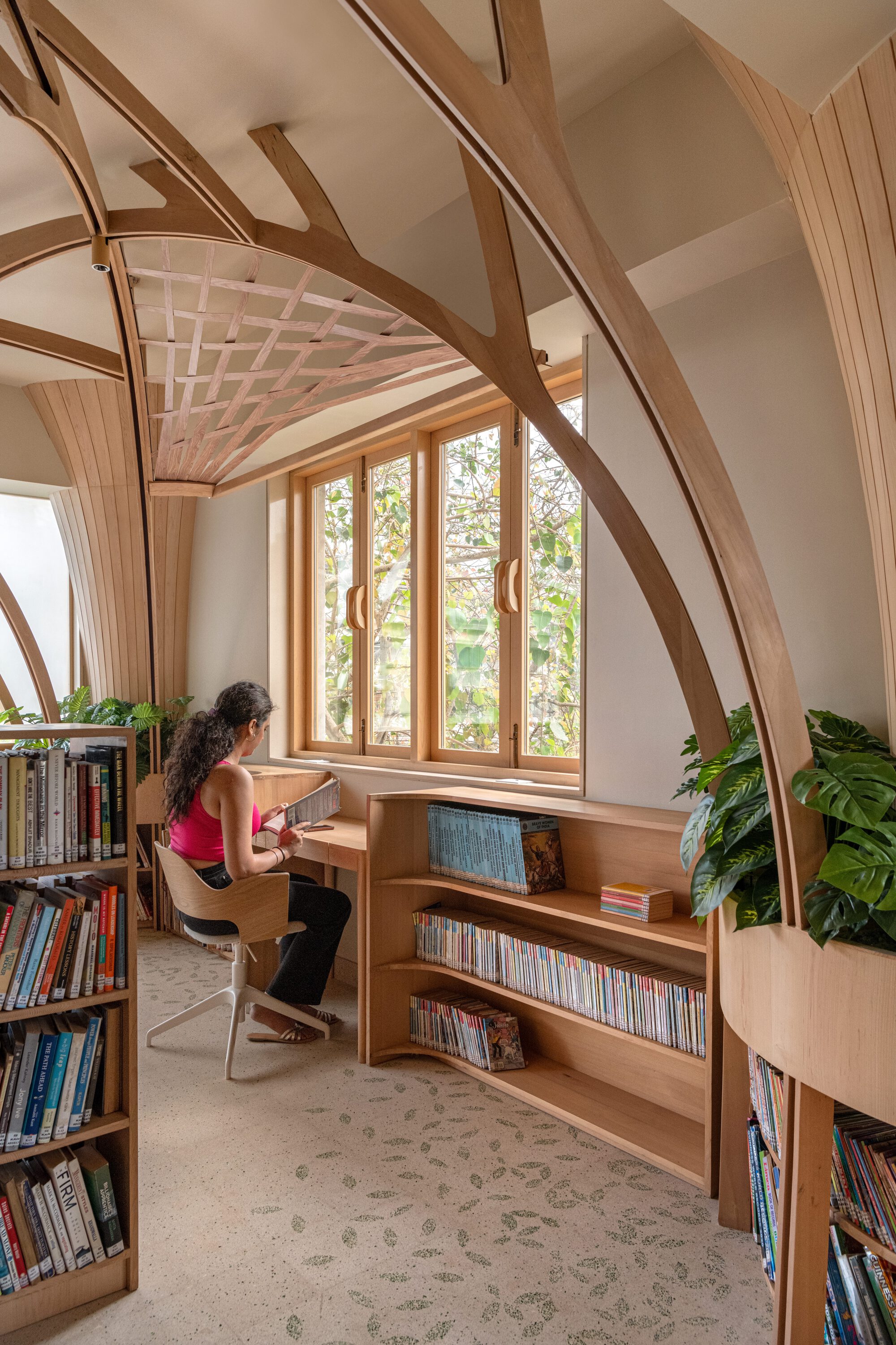Woman reading at a built-in desk under a timber arch, with a lattice ceiling panel and leafy trees outside the window