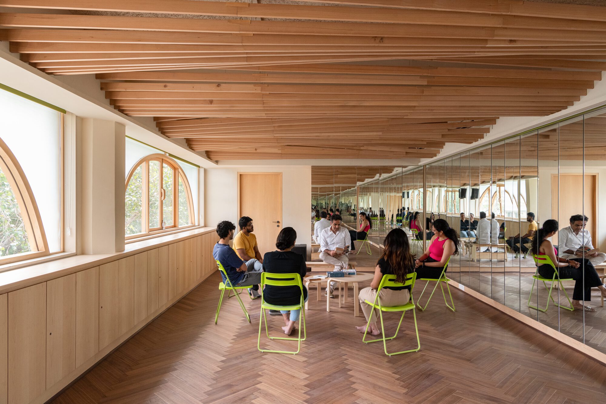 Reading room with herringbone timber floor, arched windows and a mirrored wall, where people sit on lime green chairs in soft daylight