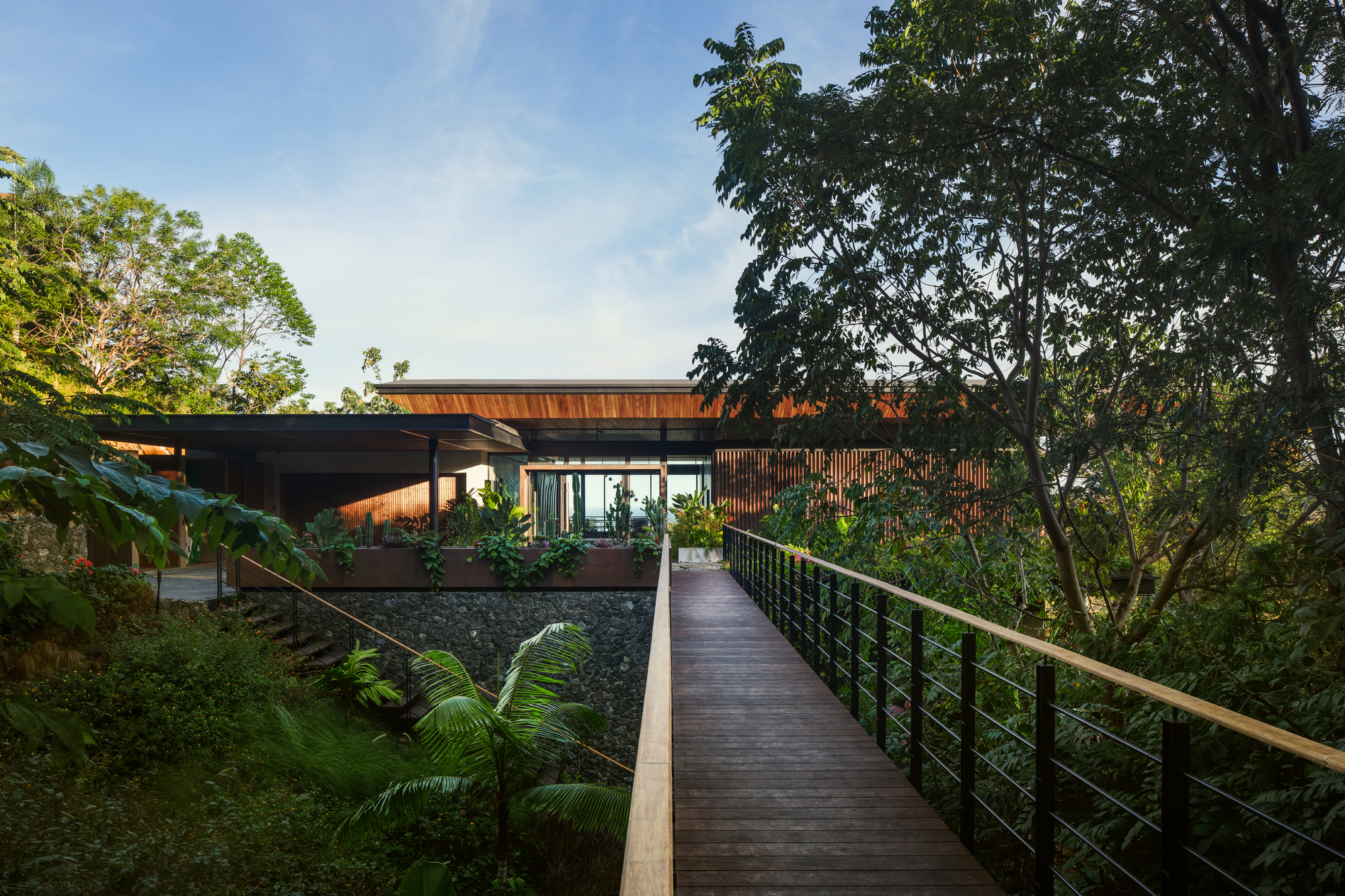 Timber boardwalk with steel cable railing leading through tropical foliage to a low pavilion with a cantilevered wood roof in late light