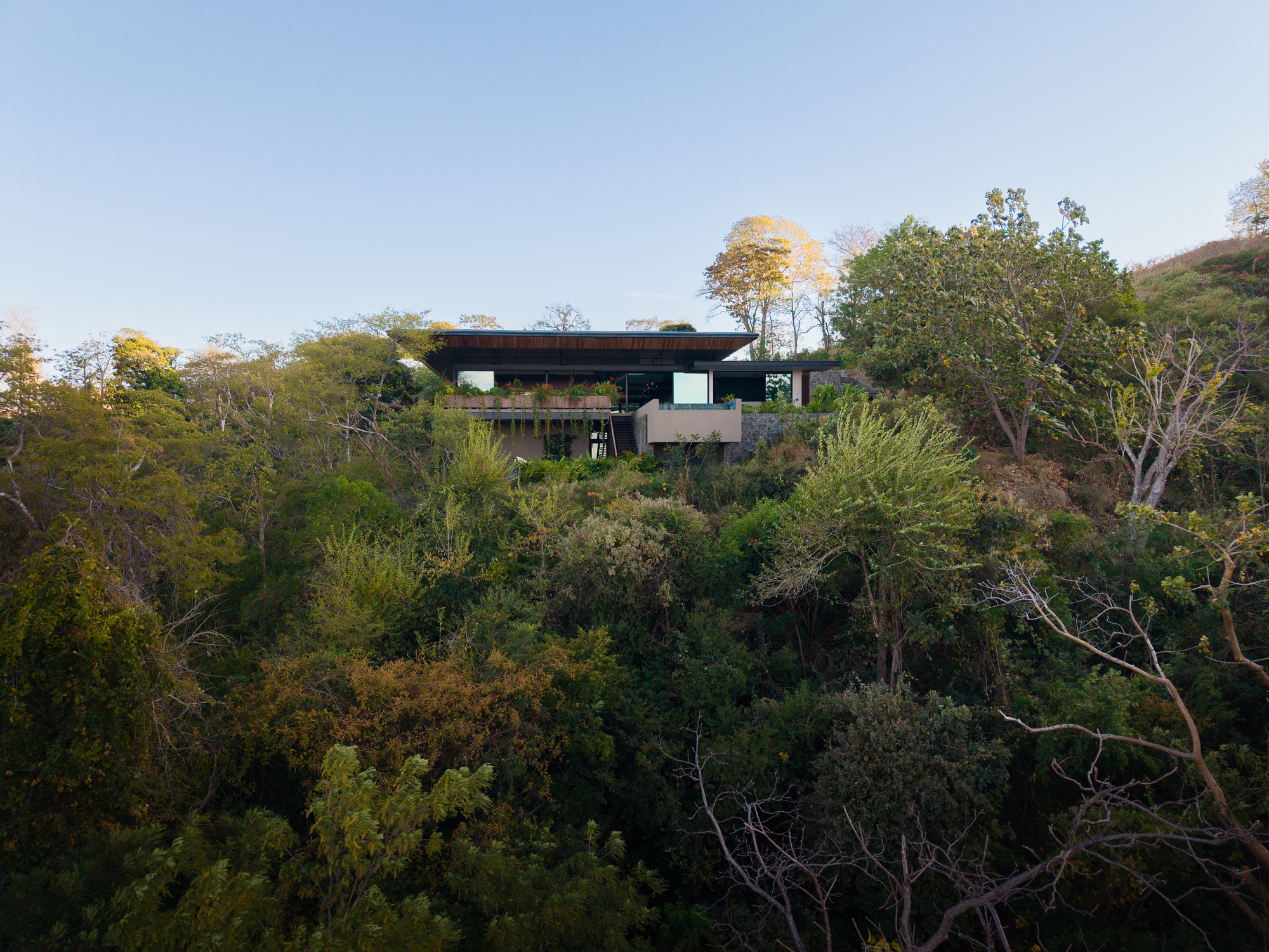 Distant view of the flat-roofed house perched on a forested hillside above dense canopy under a clear dusk sky