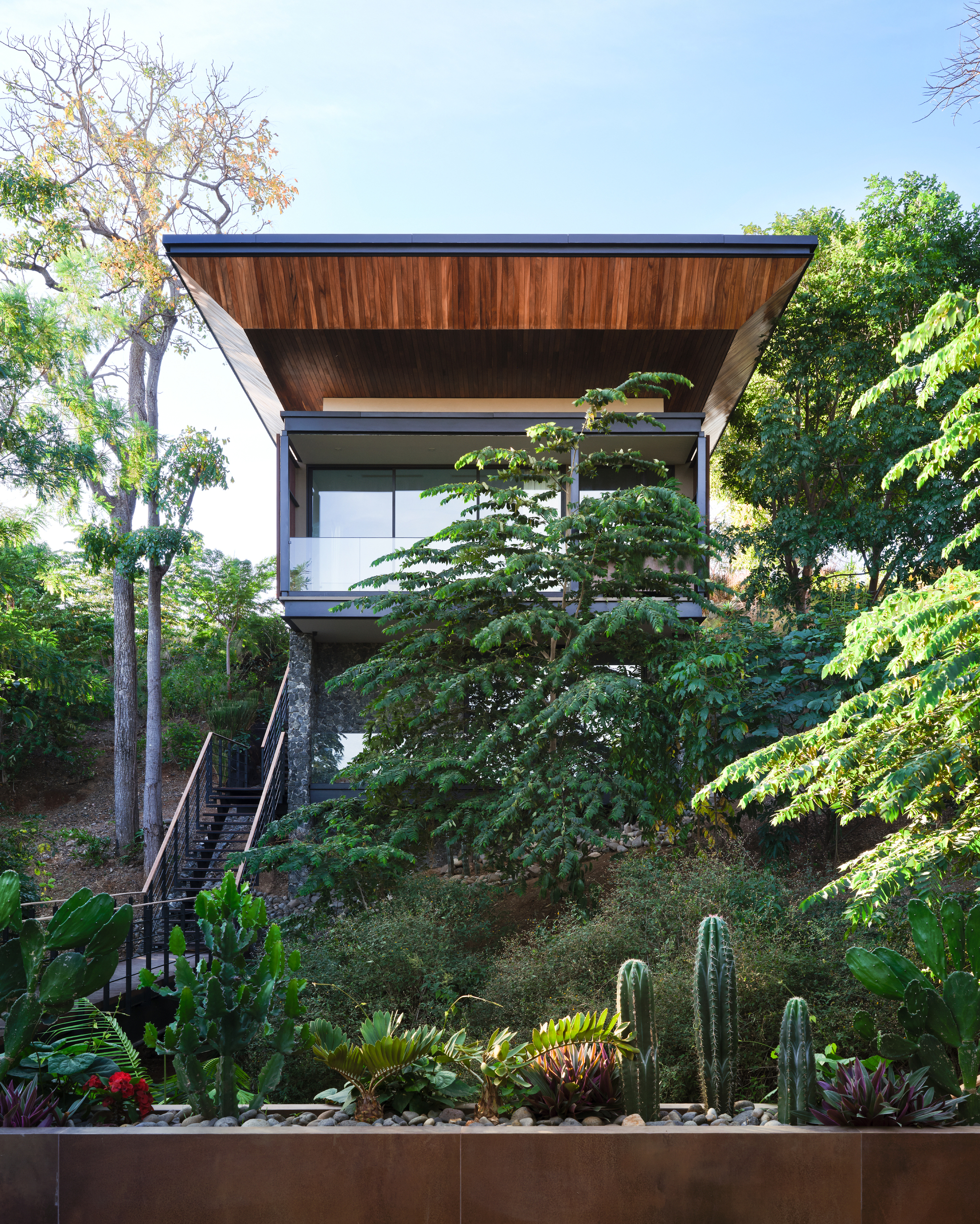 Front view of the raised house with a sloped wood-lined roof, stone pier and stair, behind cacti and a weathering steel planter