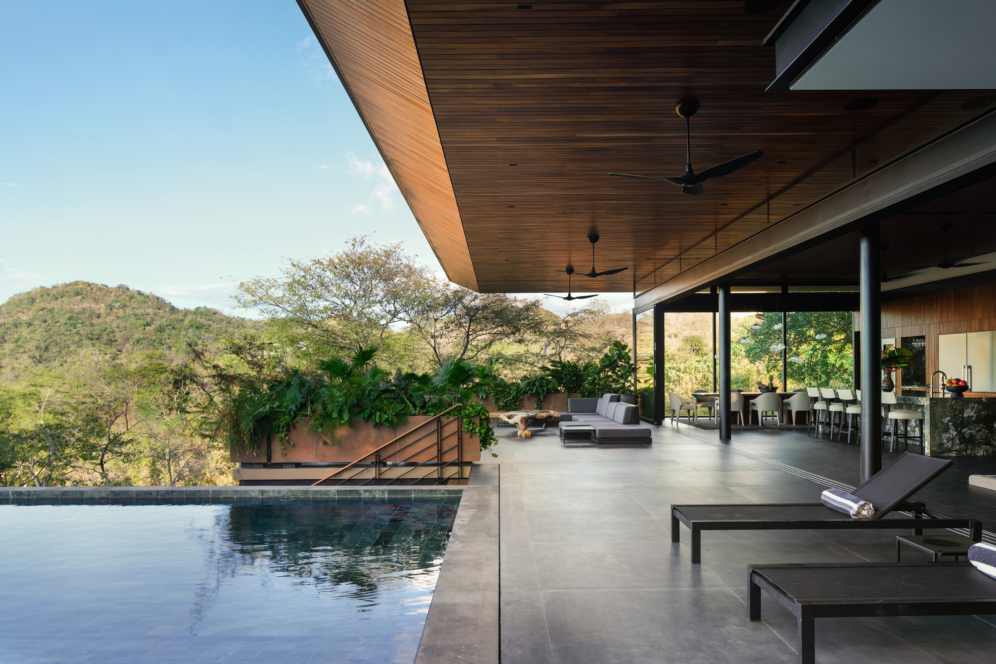 Covered terrace beside an infinity pool, with ceiling fans, lounge seating and open kitchen under a wood ceiling, hills beyond in clear daylight