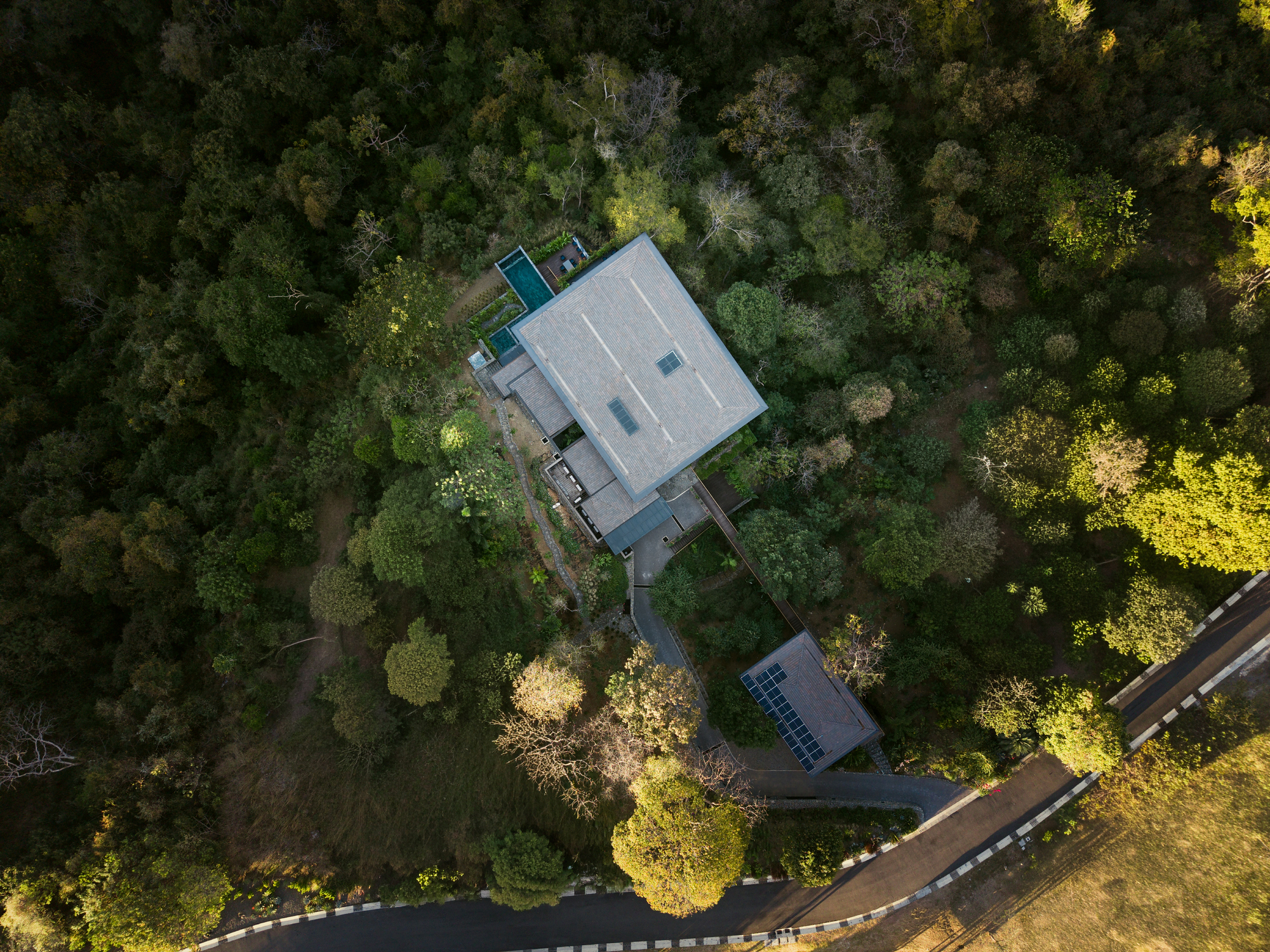 Aerial view of the flat-roofed house and its solar-panel outbuilding, set among dense forest beside a curving road