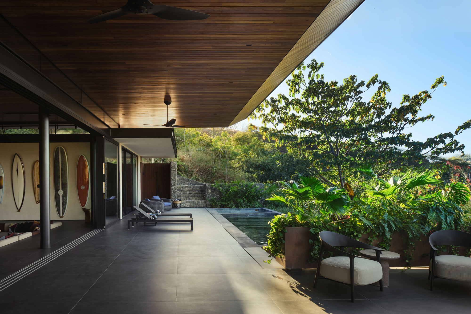 Covered terrace with timber ceiling, surfboards on the wall and a plunge pool edged by planters in afternoon sun
