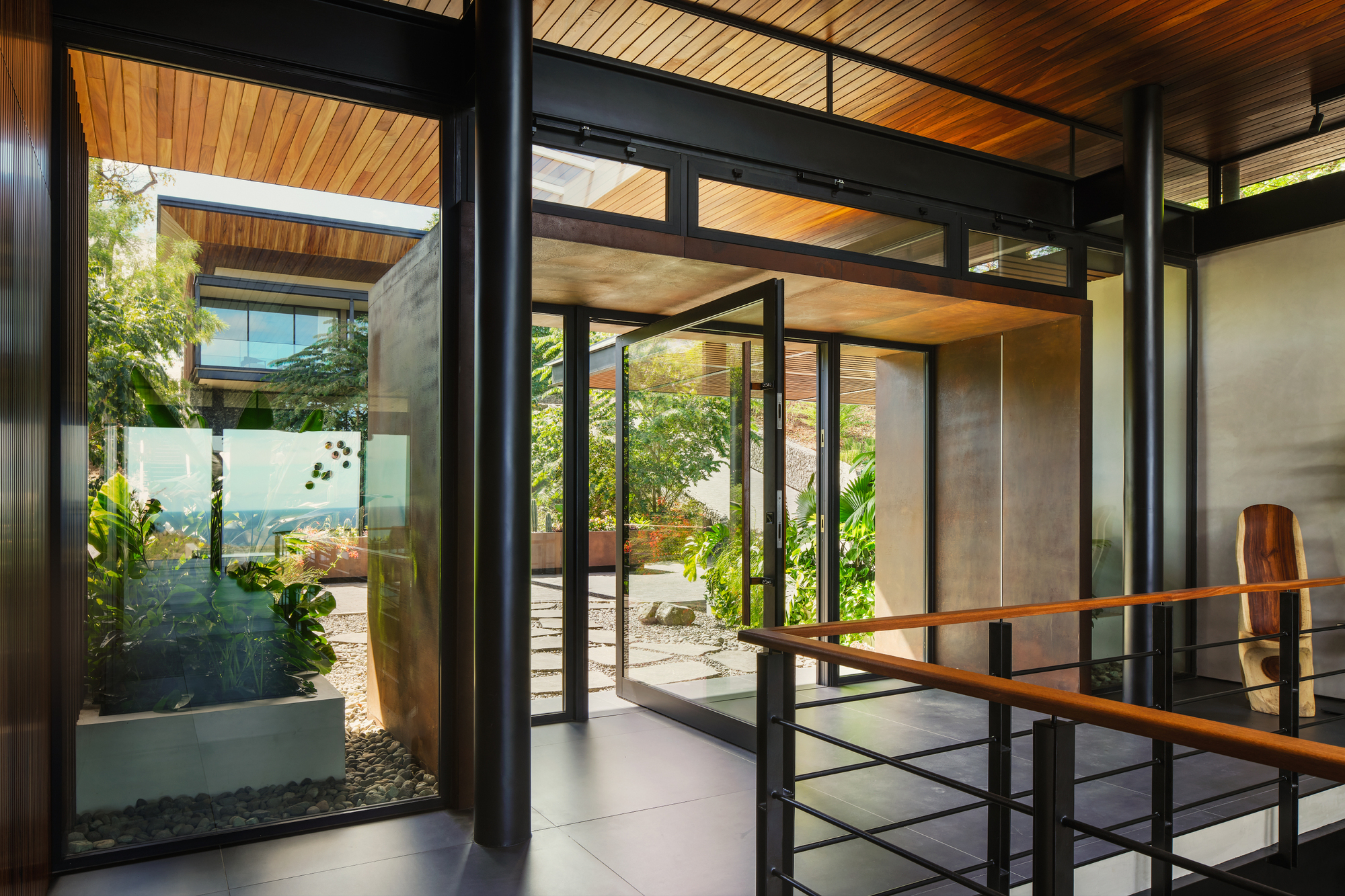 Entry hall with black steel columns, a pivot glass door and a planted gravel courtyard glimpsed through the glazing